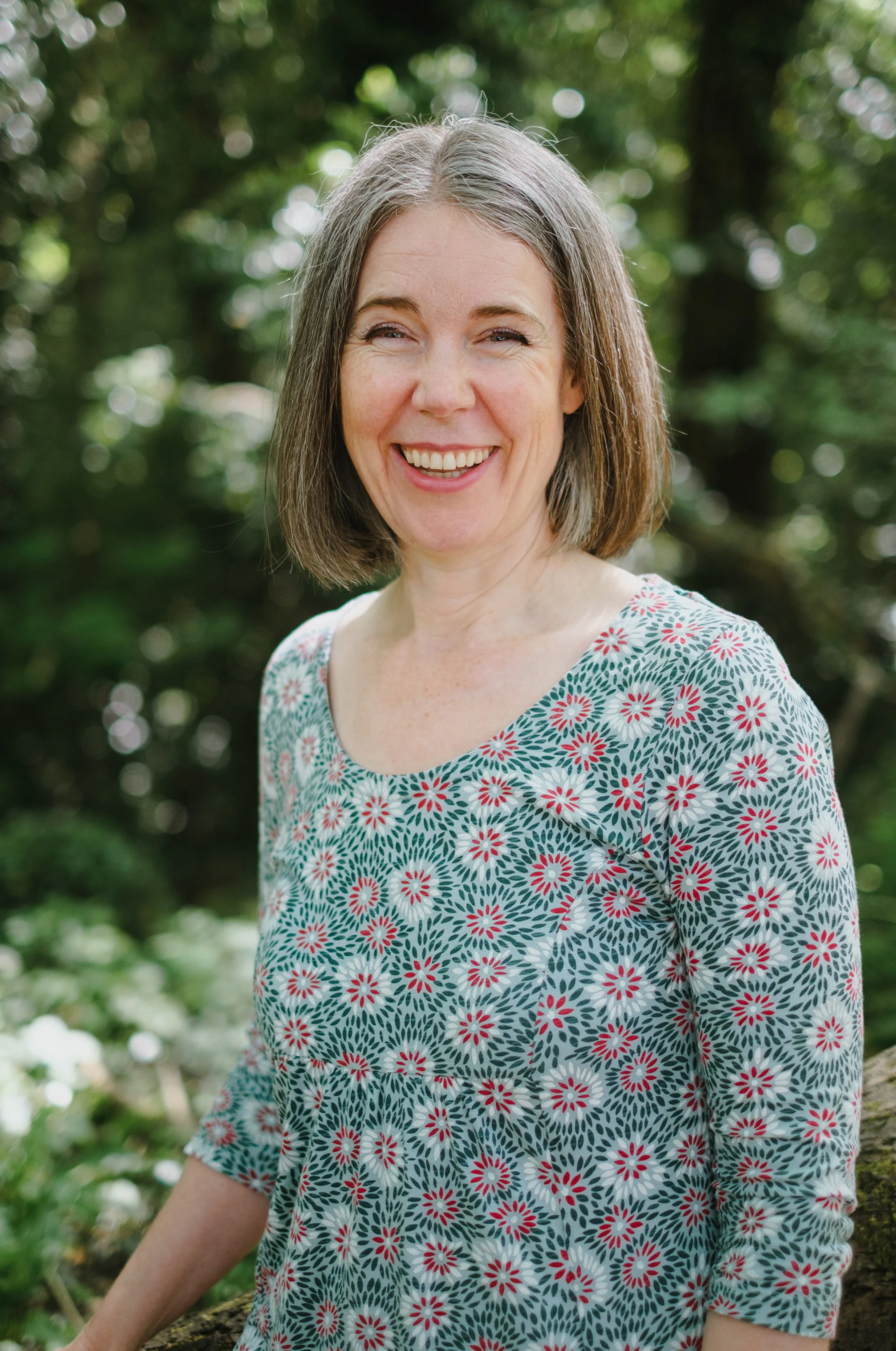 A woman with shoulder-length gray hair smiling outdoors in a green, leafy area, wearing a patterned dress with white, pink, and green colors.