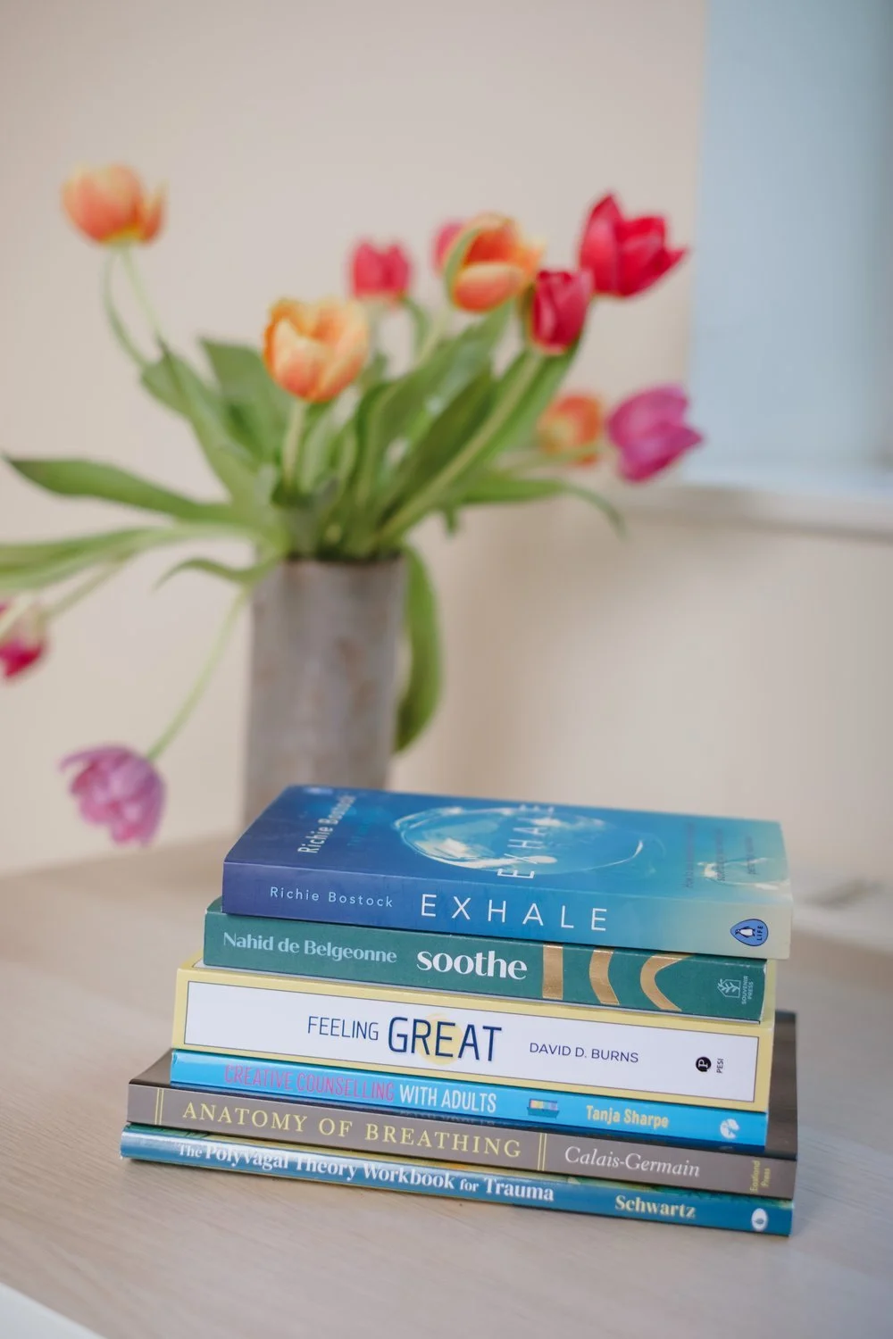 A stack of therapy and wellbeing books on a table with a vase of colourful tulips in the background