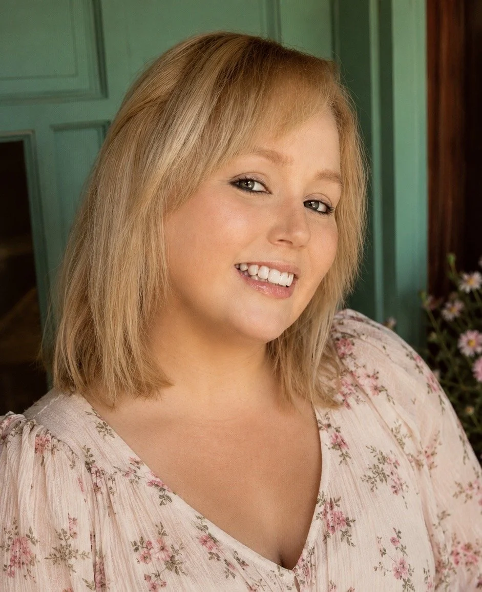 A young woman with blonde hair smiling, wearing a floral dress, standing in front of green paneled walls with flowers on the side.