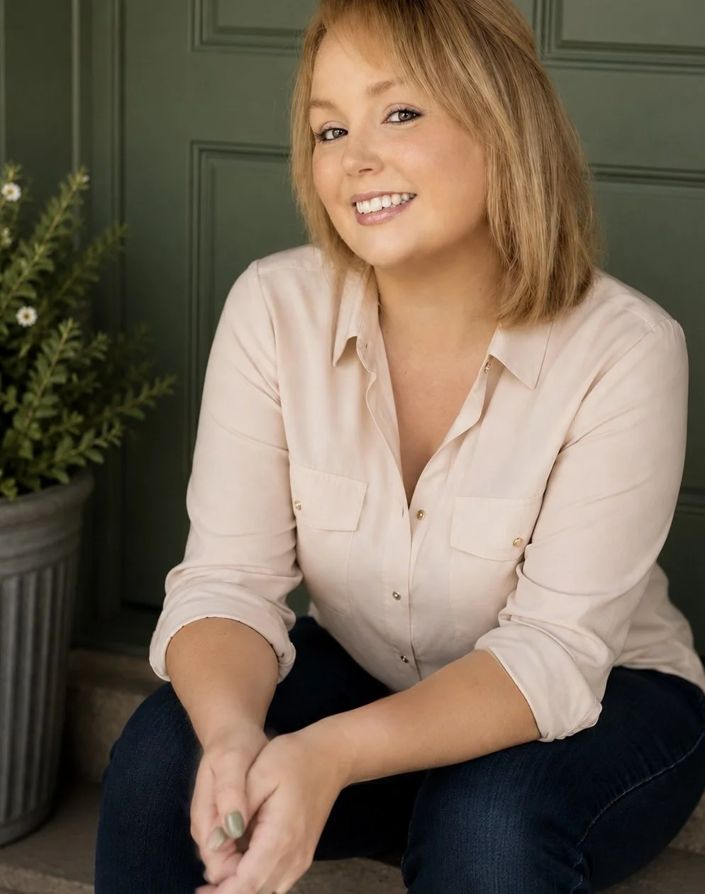 A woman with shoulder-length blond hair sitting in front of a green wall and a potted plant, smiling at the camera wearing a light-colored blouse and dark jeans.