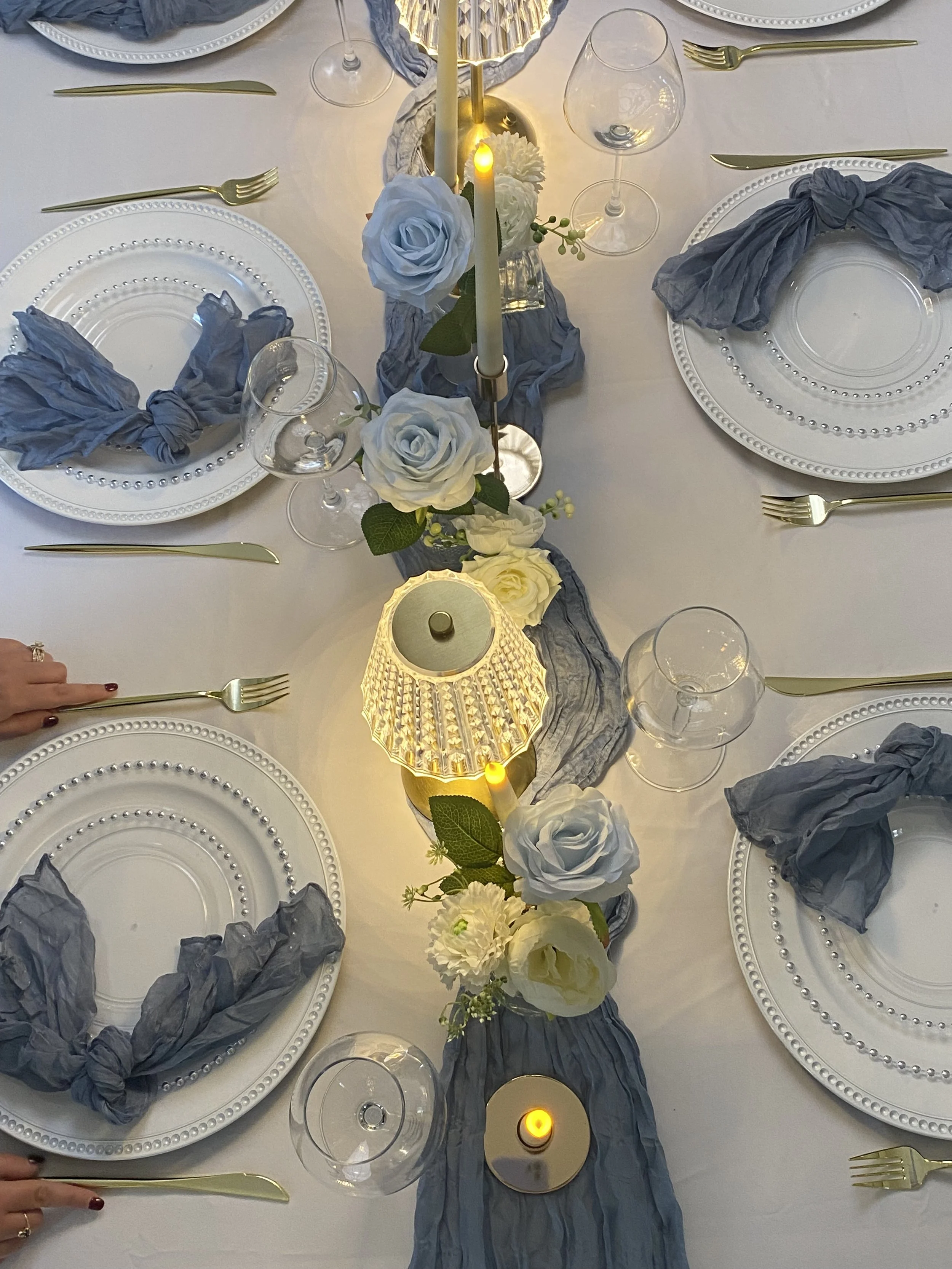 A formal dinner table setting with white plates, gold utensils, wine glasses, and a floral centerpiece with white roses, white flowers, and greenery, accented by a gray and white fabric table runner and small candles.