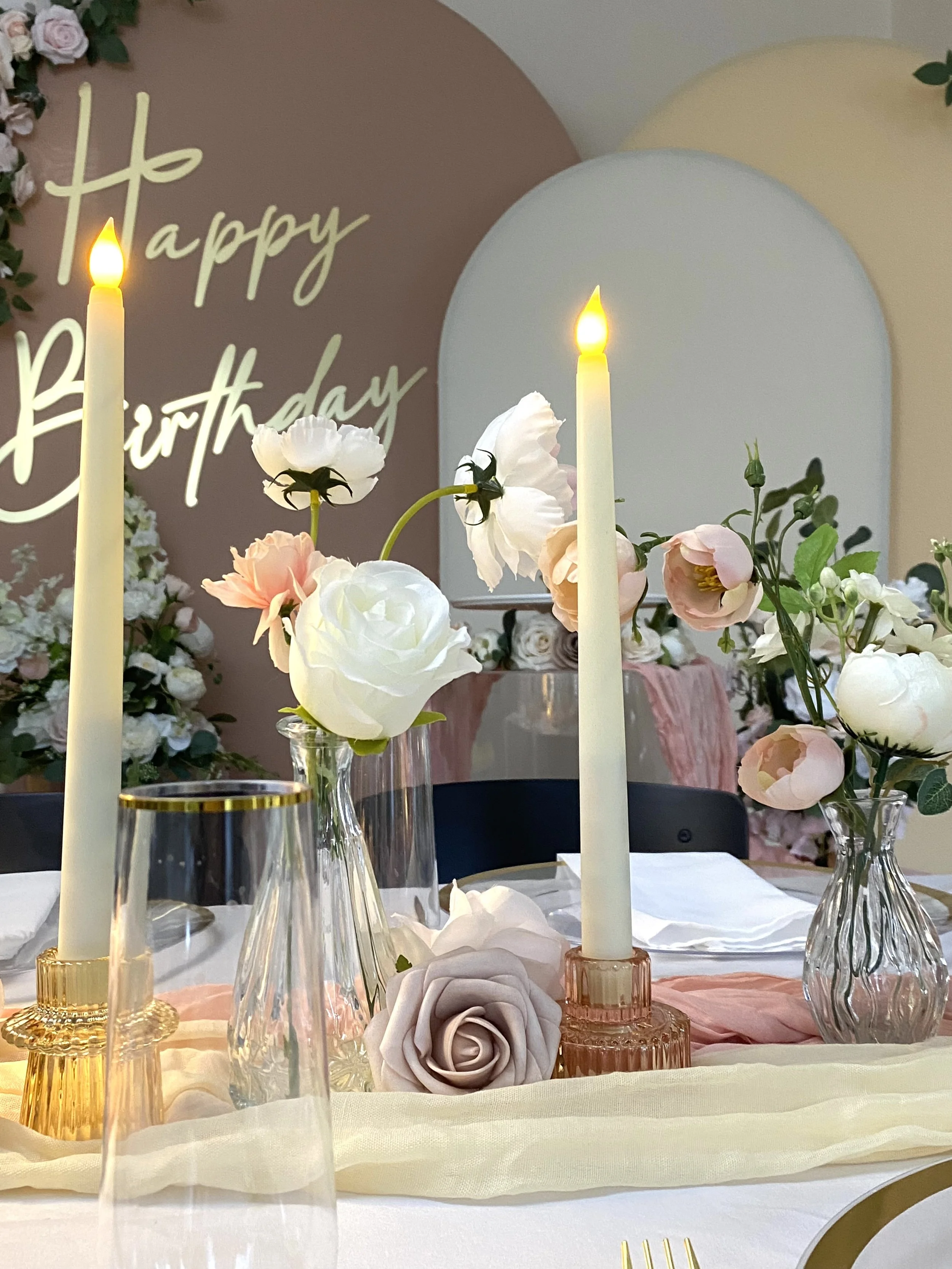 Elegant birthday celebration table decorated with pink, white, and peach flowers, tall candles, glass vases, and a 'Happy Birthday' sign in the background.