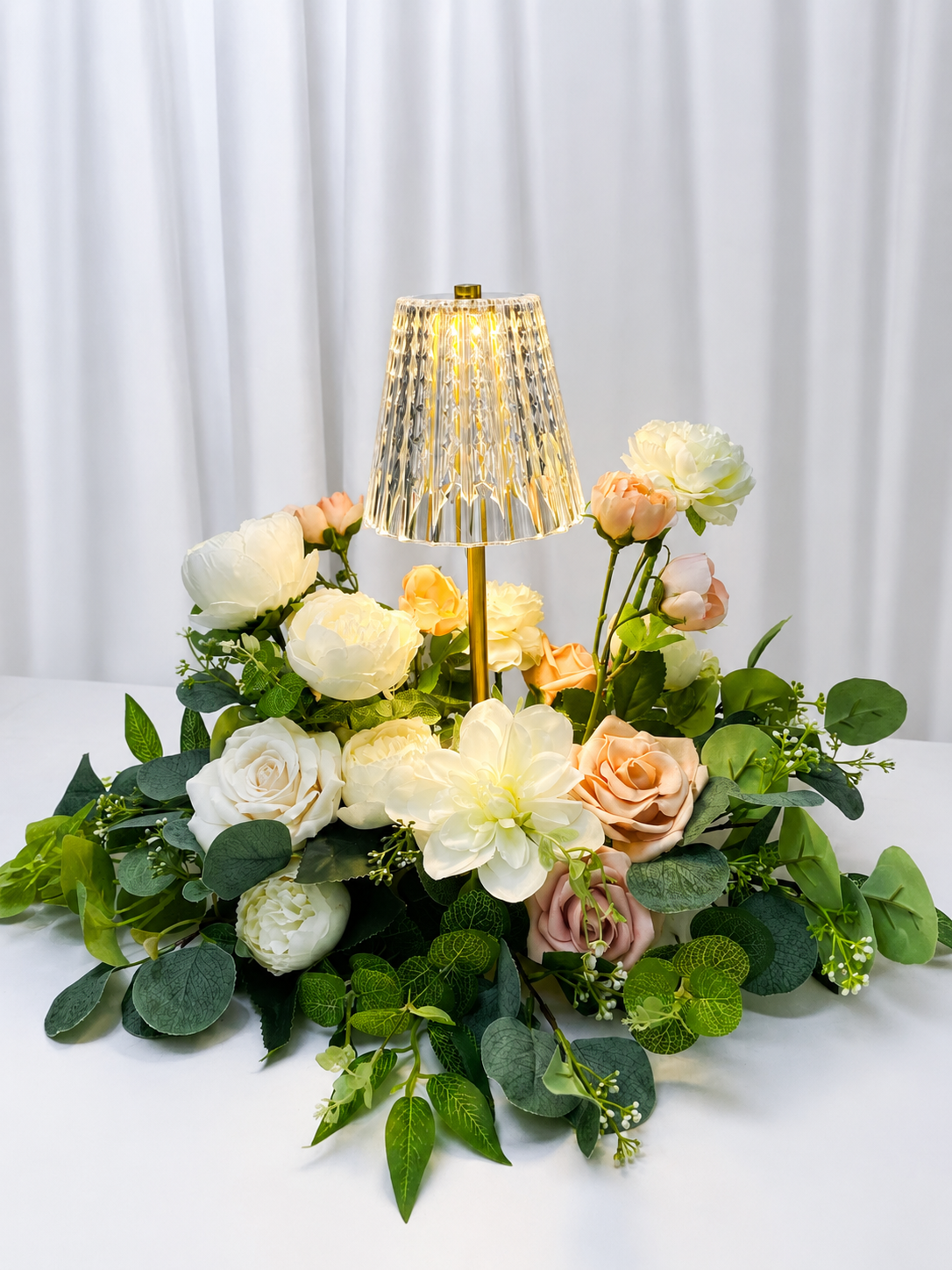 A tabletop floral arrangement with white, cream, and peach roses, topped with a small gold and crystal table lamp, set against a white curtain backdrop.