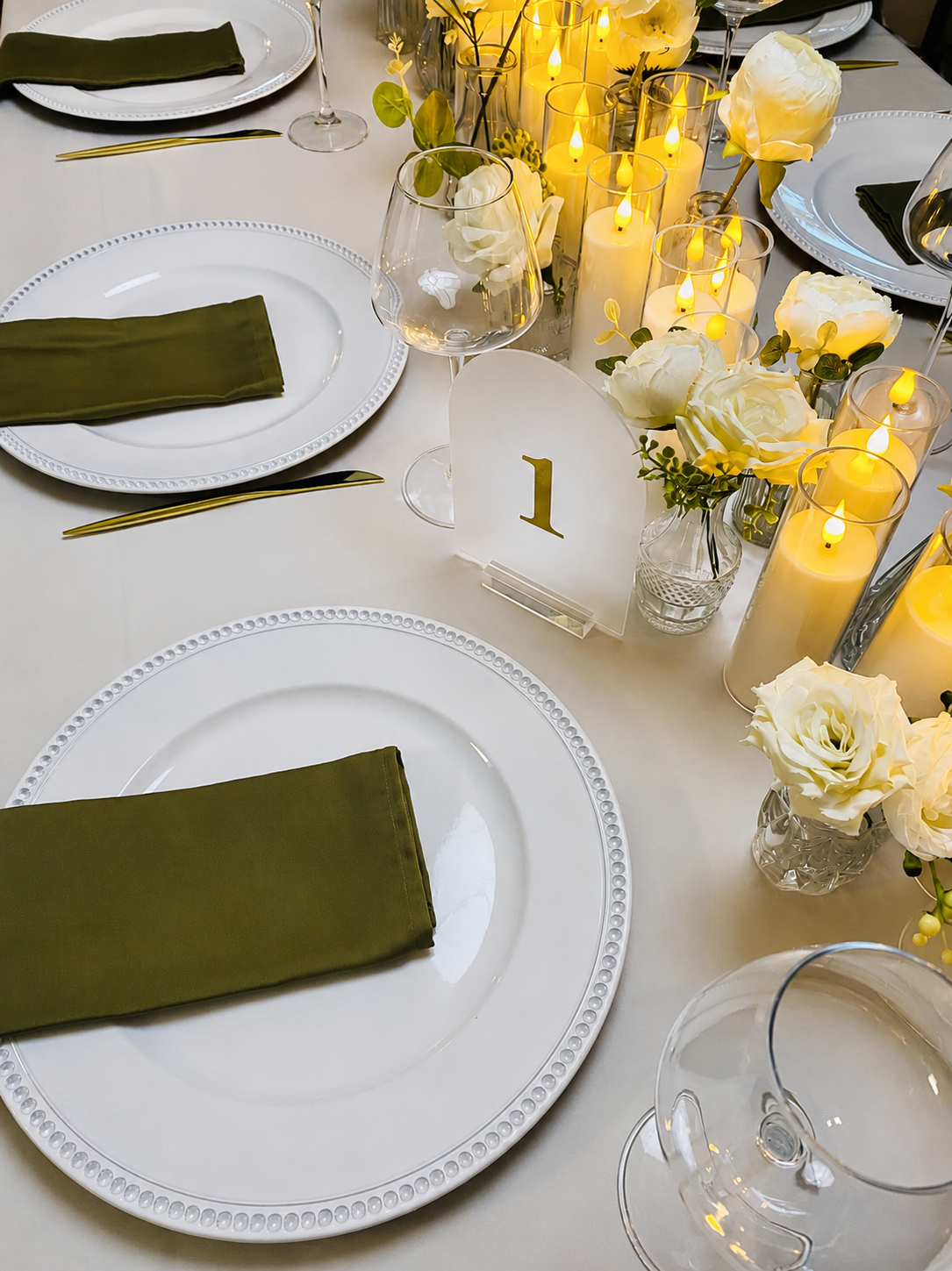Elegant dinner table setup with white plates, green napkins, gold utensils, wine glasses, candles, and white floral centerpieces, featuring a table number card with the number one.