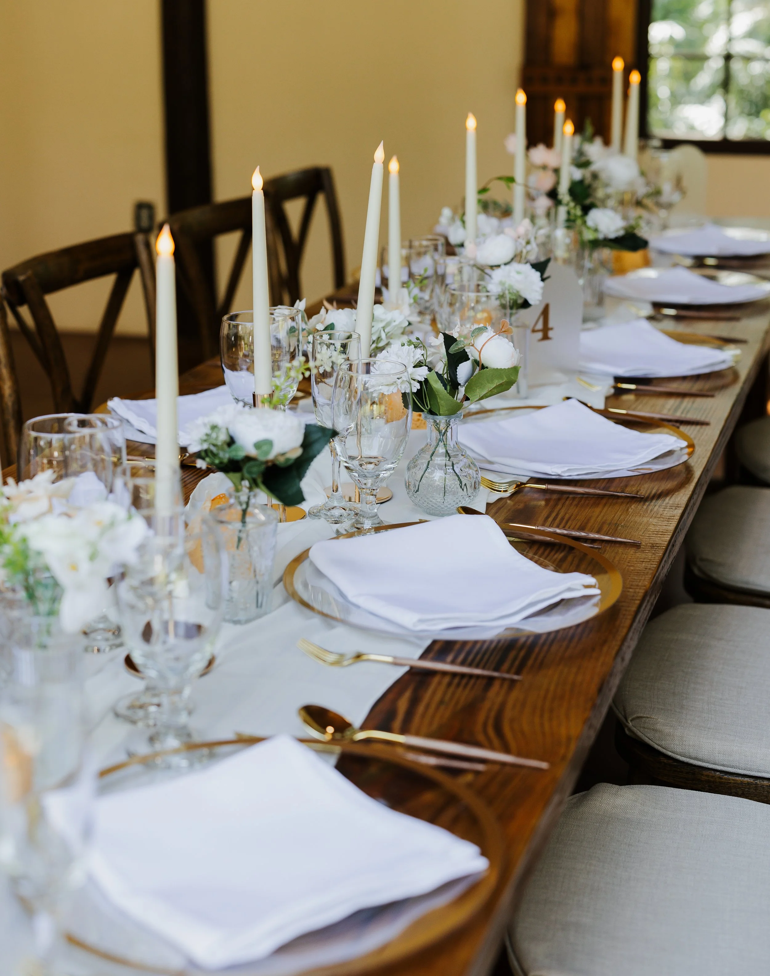 A decorated dining table with white candles, floral arrangements, glassware, plates, and cutlery, set for a formal event.