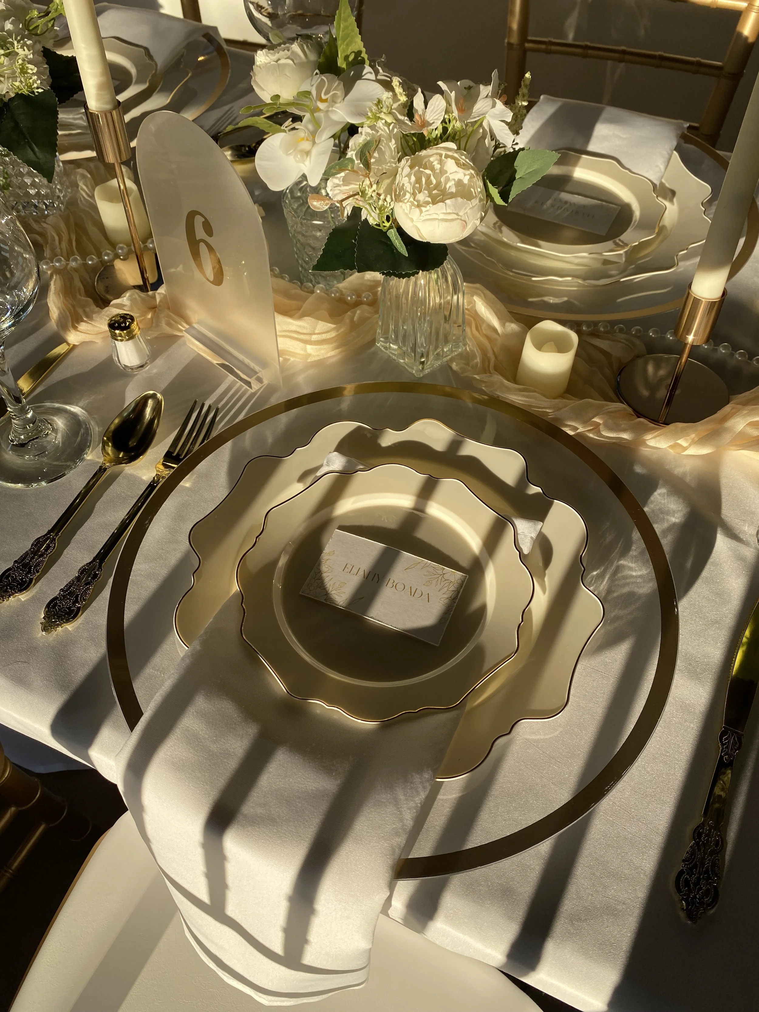 Elegant table setting with layered gold-rimmed plates, a floral centerpiece, candles, pearls, and a place card reading 'Helen Bodi.'