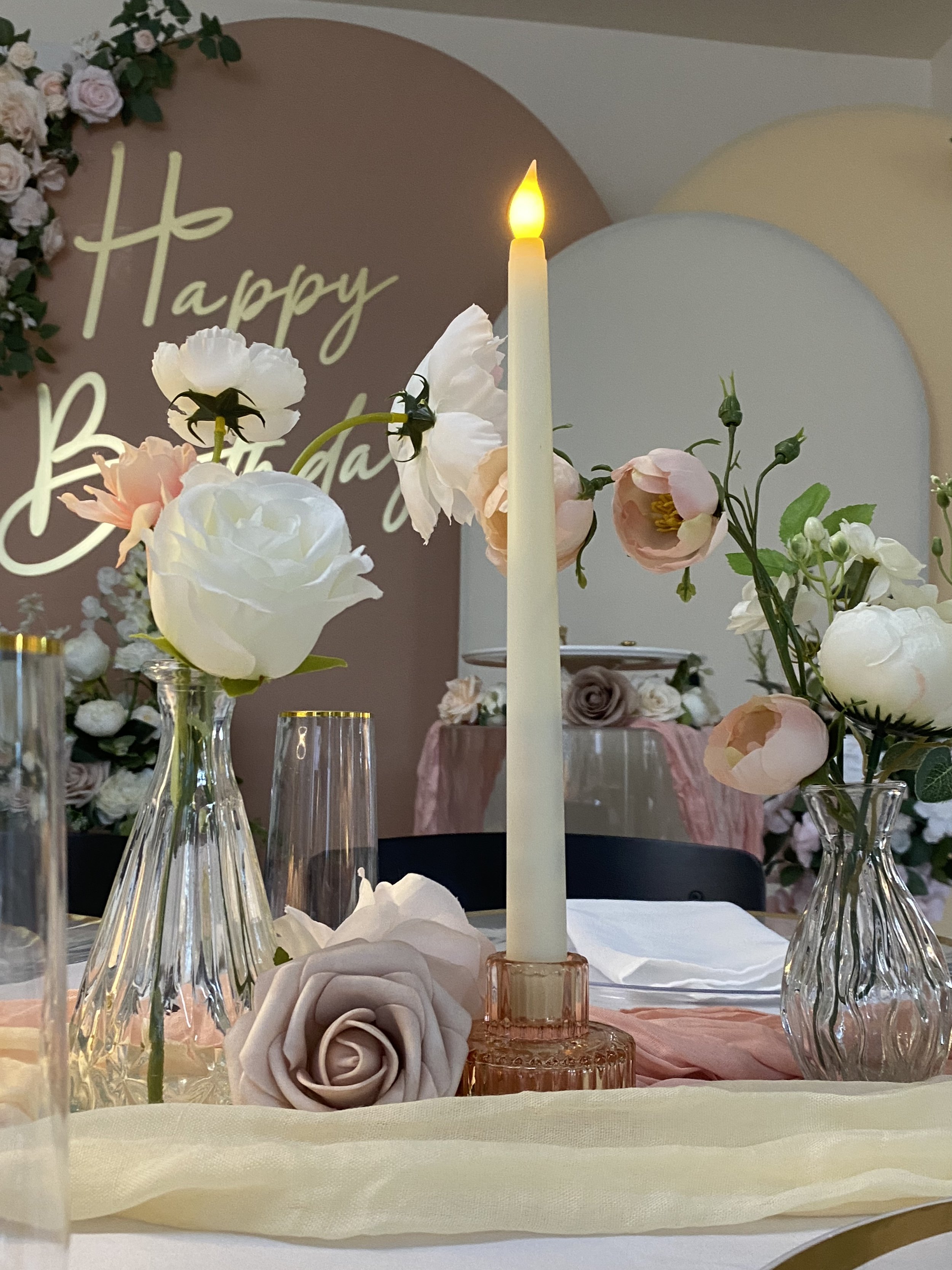 A table decorated for a celebration with flowers in glass vases, a lit candle, a pink rose-shaped decoration, and a backdrop with the words 'Happy Birthday' and floral decorations.
