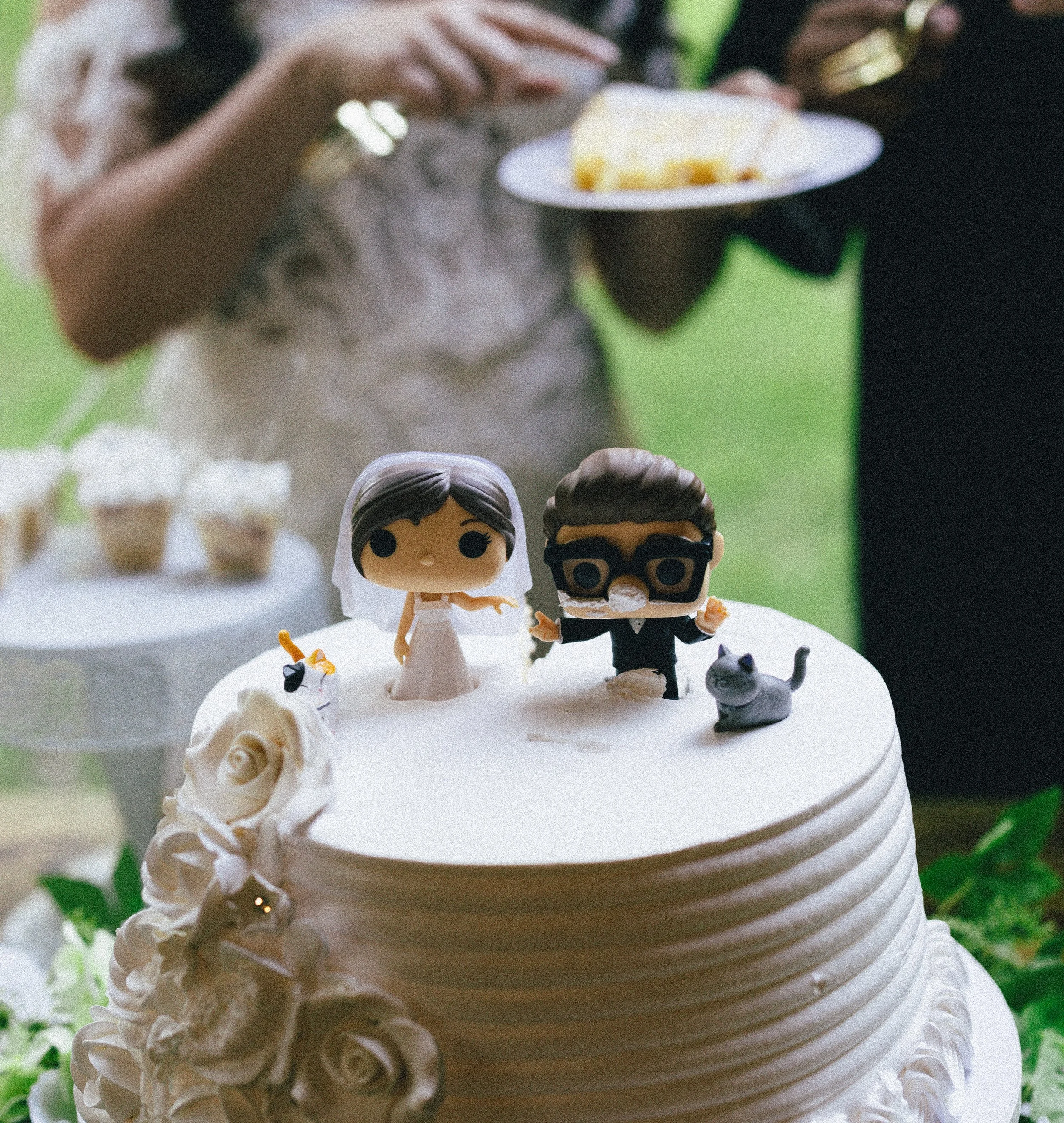 Close-up of a wedding cake with white icing, decorated with cream-colored roses and small figurines of a bride and groom on the top tier, set against a blurred green background.