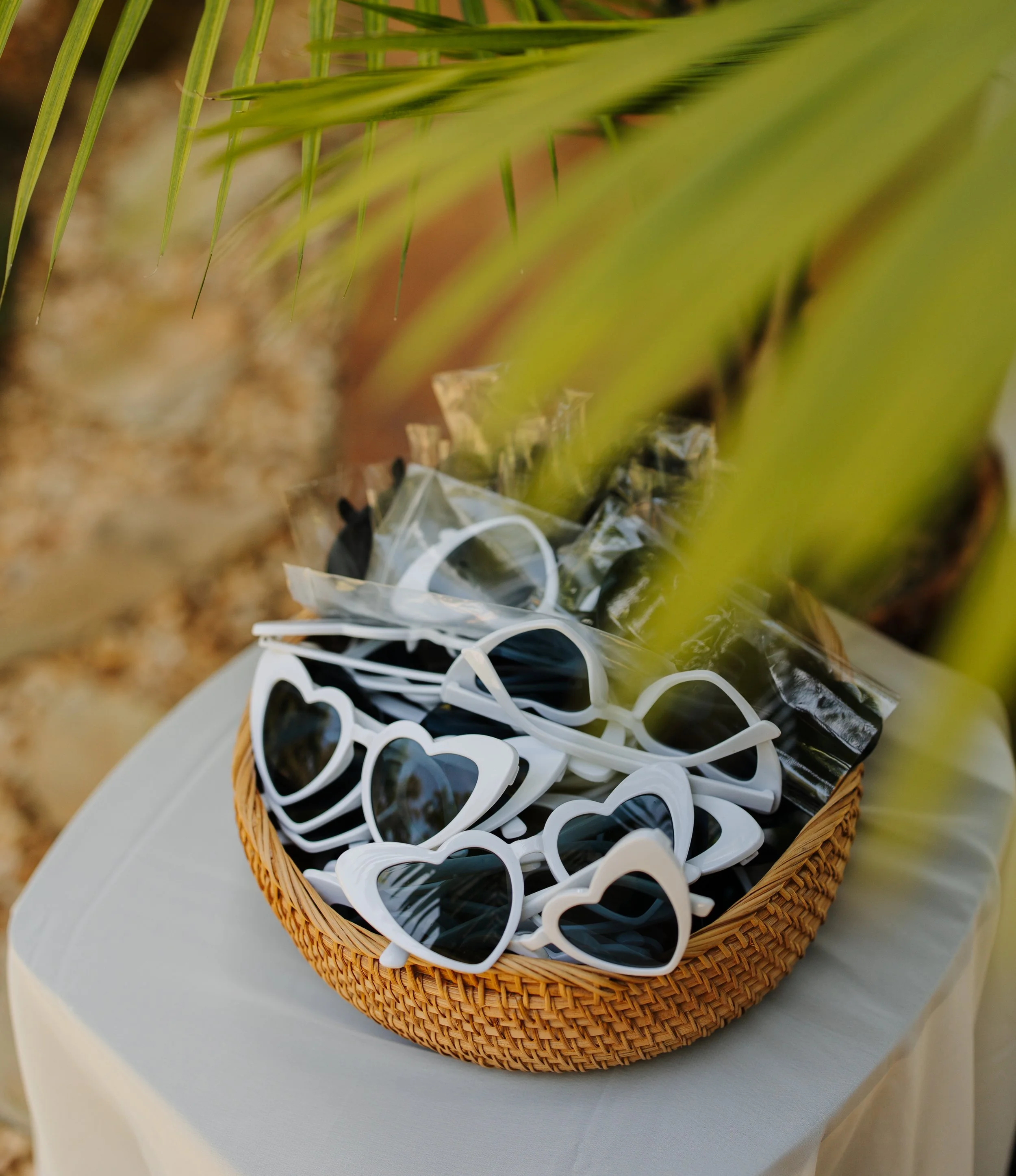 A basket full of heart-shaped sunglasses with dark lenses and white frames, placed on a white table.