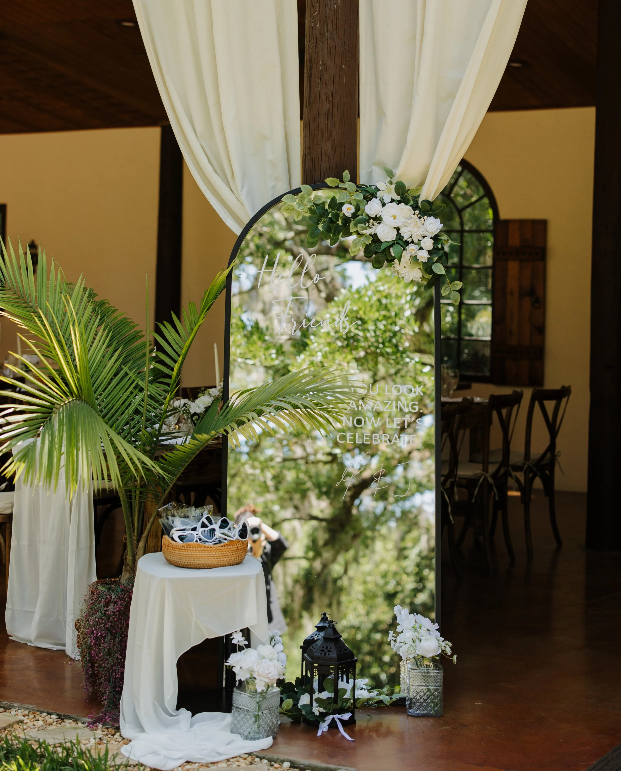 Decorative wedding or event setup with white flowers, greenery, lanterns, and a sign that reads "Hello friends. You look amazing. Now let's celebrate!". The backdrop features cream curtains and a wooden beam, with natural light and greenery visible outside.
