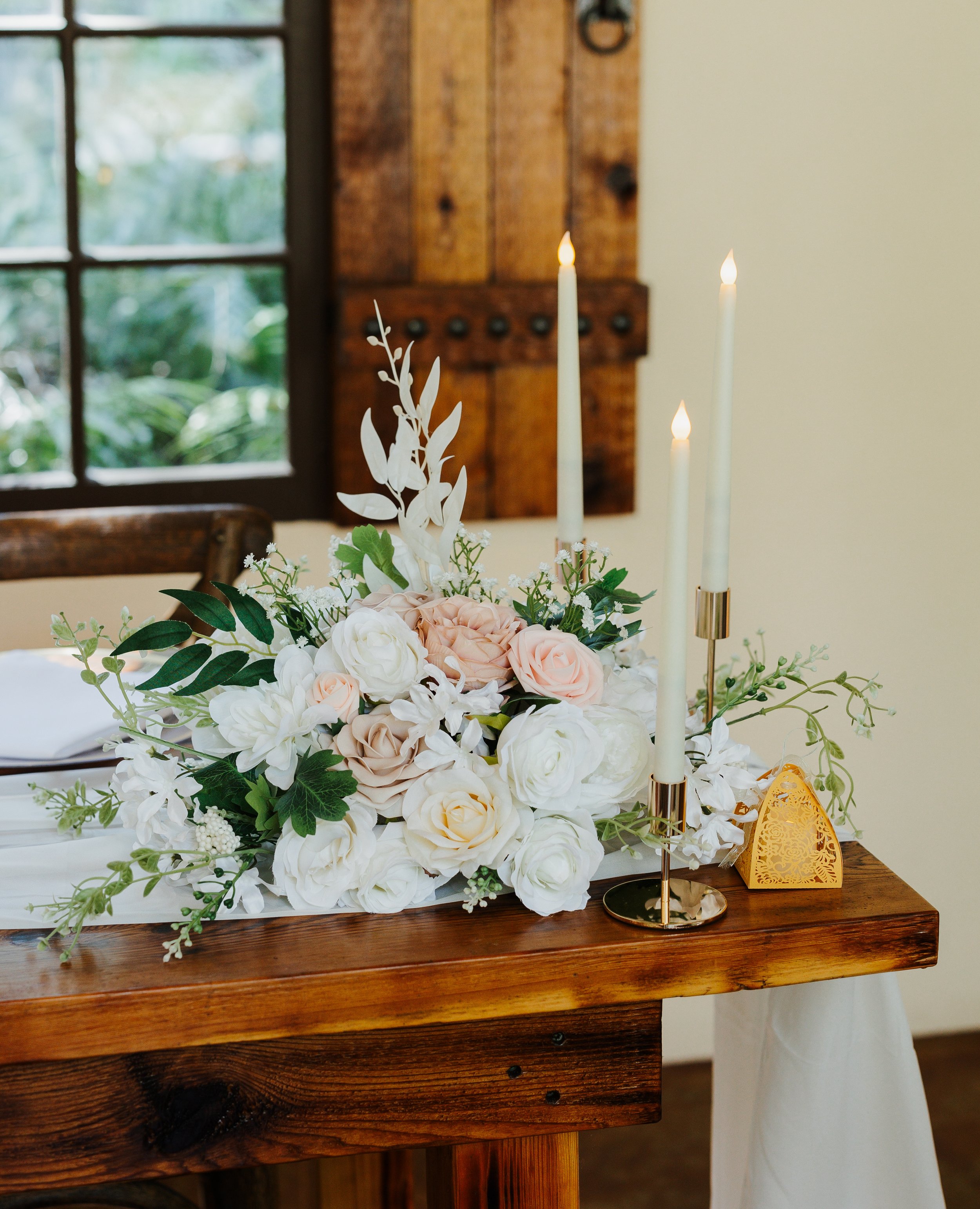 A floral centerpiece with white and blush roses, white peonies, and green foliage on a wooden table. There are three tall white candles in gold holders, and a decorative orange lattice object nearby. A window with a wooden frame and a beige wall are in the background.