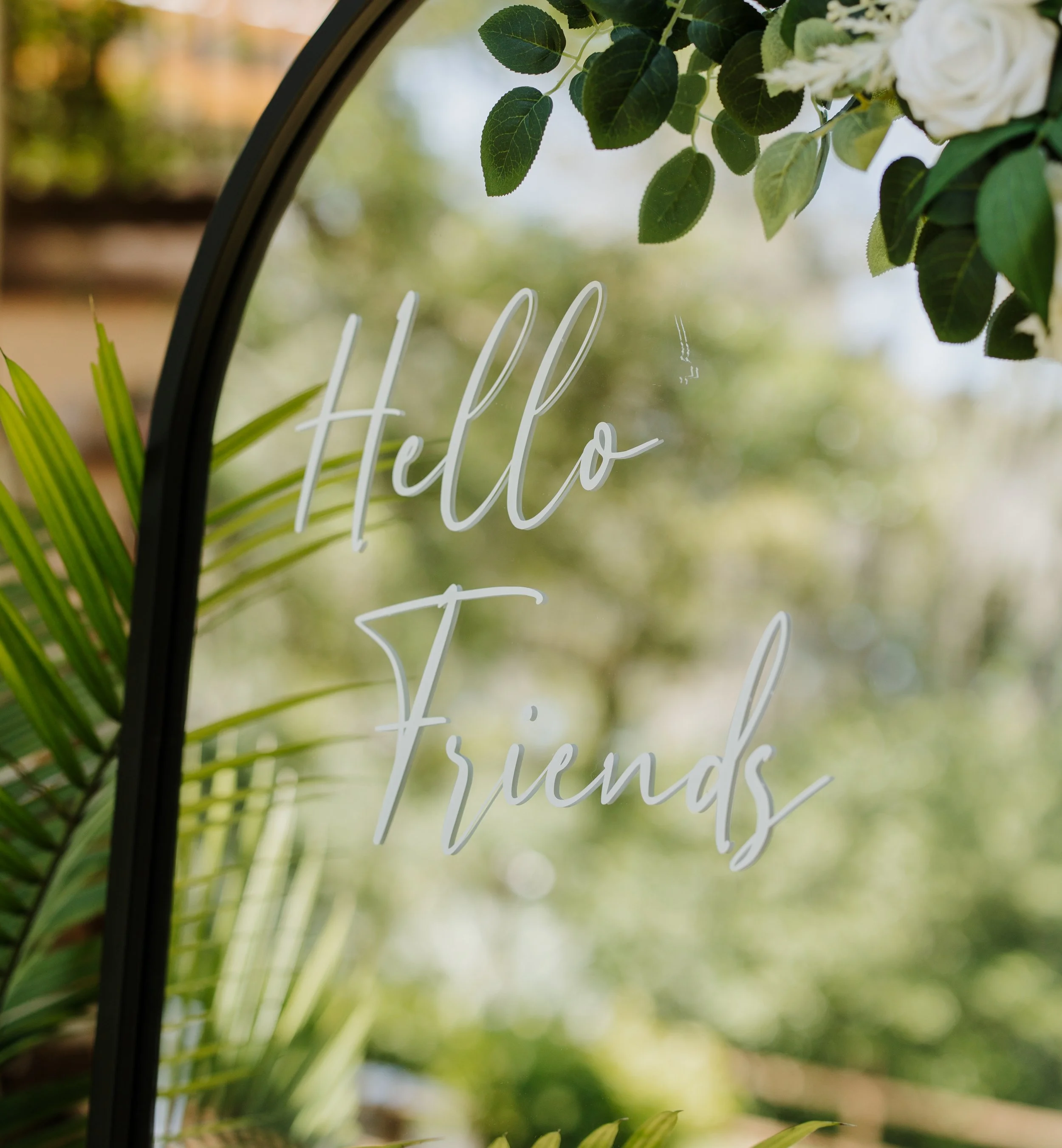 Close-up of a mirror with the words 'Hello friends' written on it, surrounded by green leaves and white flowers, with a blurred natural background.