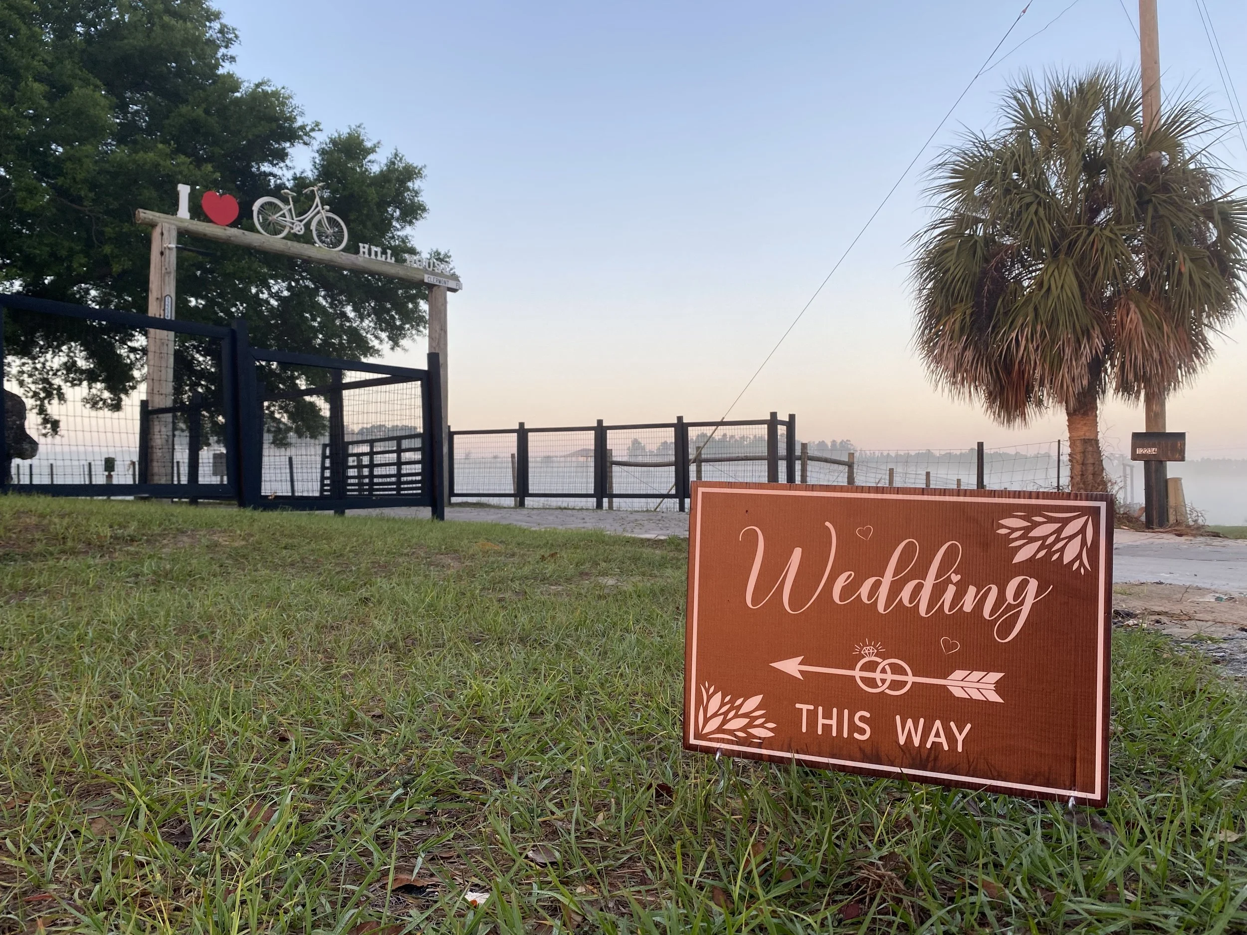 A sign with the words "Welcome to this way" and an arrow, placed on a grassy area near a wedding venue. In the background, a sign says "I love bicycle" with a red heart and a bicycle icon, and signs indicating the location as a wedding venue near a body of water, with trees and a clear sky.