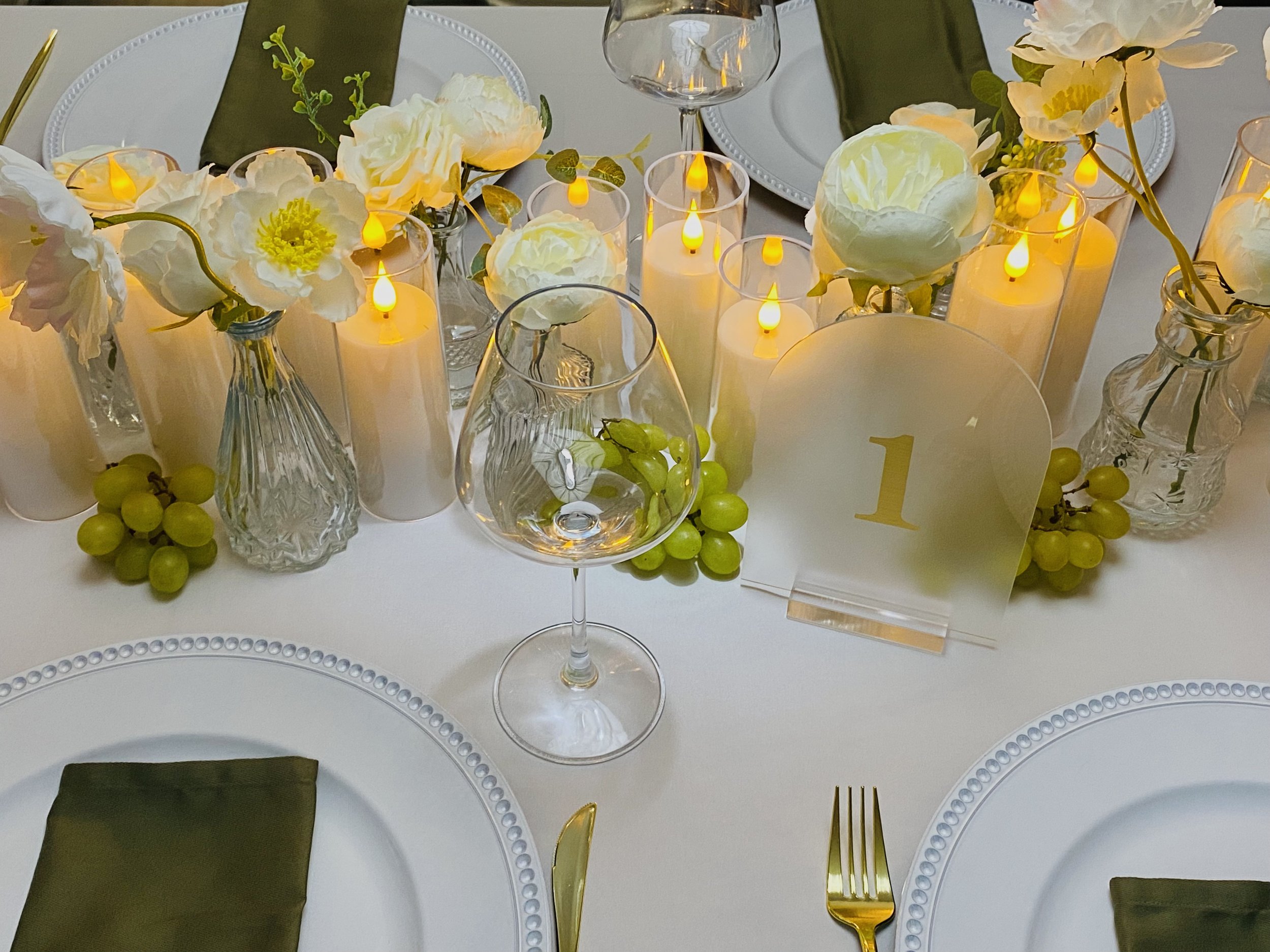 A table decorated with white flowers, green grapes, and yellow LED candles in glass holders. The table is set with white plates, napkins, and gold utensils, featuring a white tablecloth and green cloth napkins.