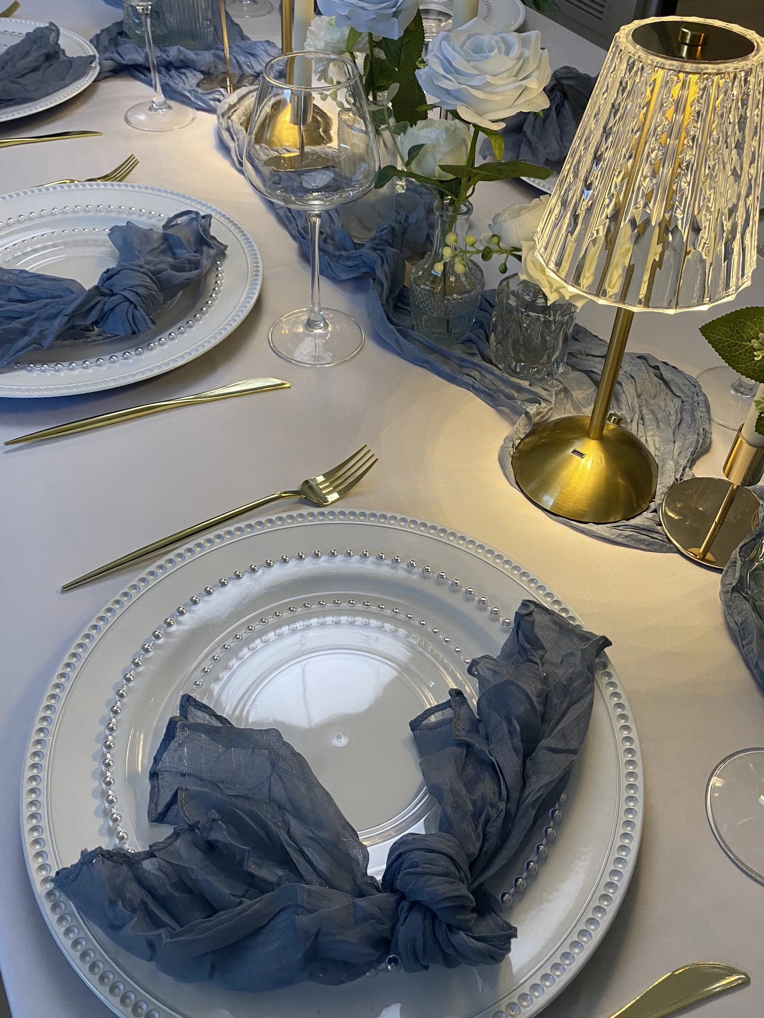 A dining table setup with white plates, golden cutlery, a blue and white color theme, a floral centerpiece with white roses and green foliage, and a small table lamp with a gold base and a pleated shade.