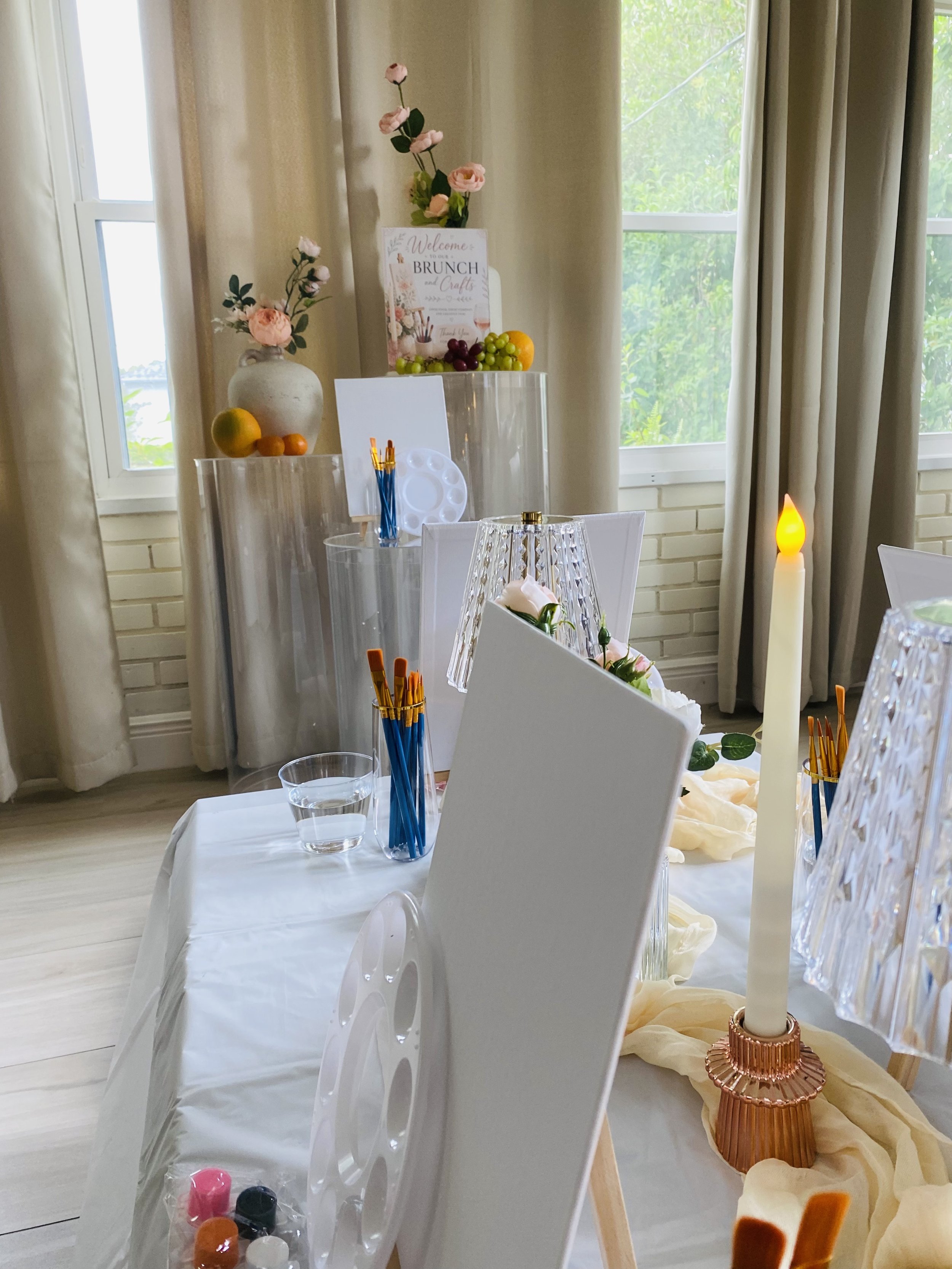 Decorative table setting with candles, flowers, and balloons in a room with large windows and beige curtains.