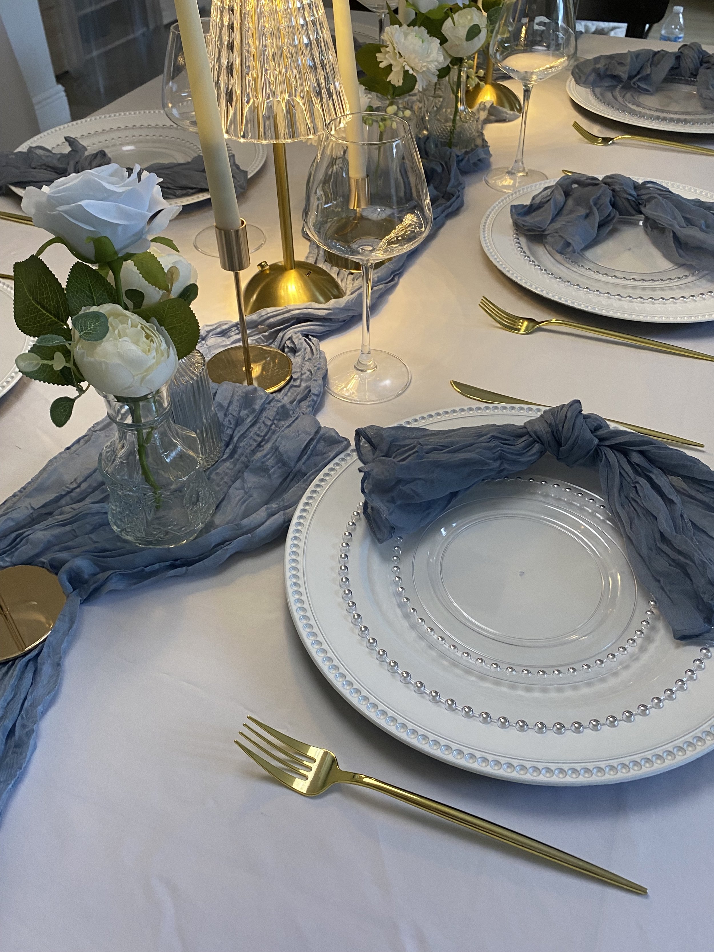 Elegant dining table set with white tablecloth, decorative plates, gold utensils, gray napkins, floral centerpiece, and candelabra with candles. Soft lighting creates a sophisticated atmosphere.