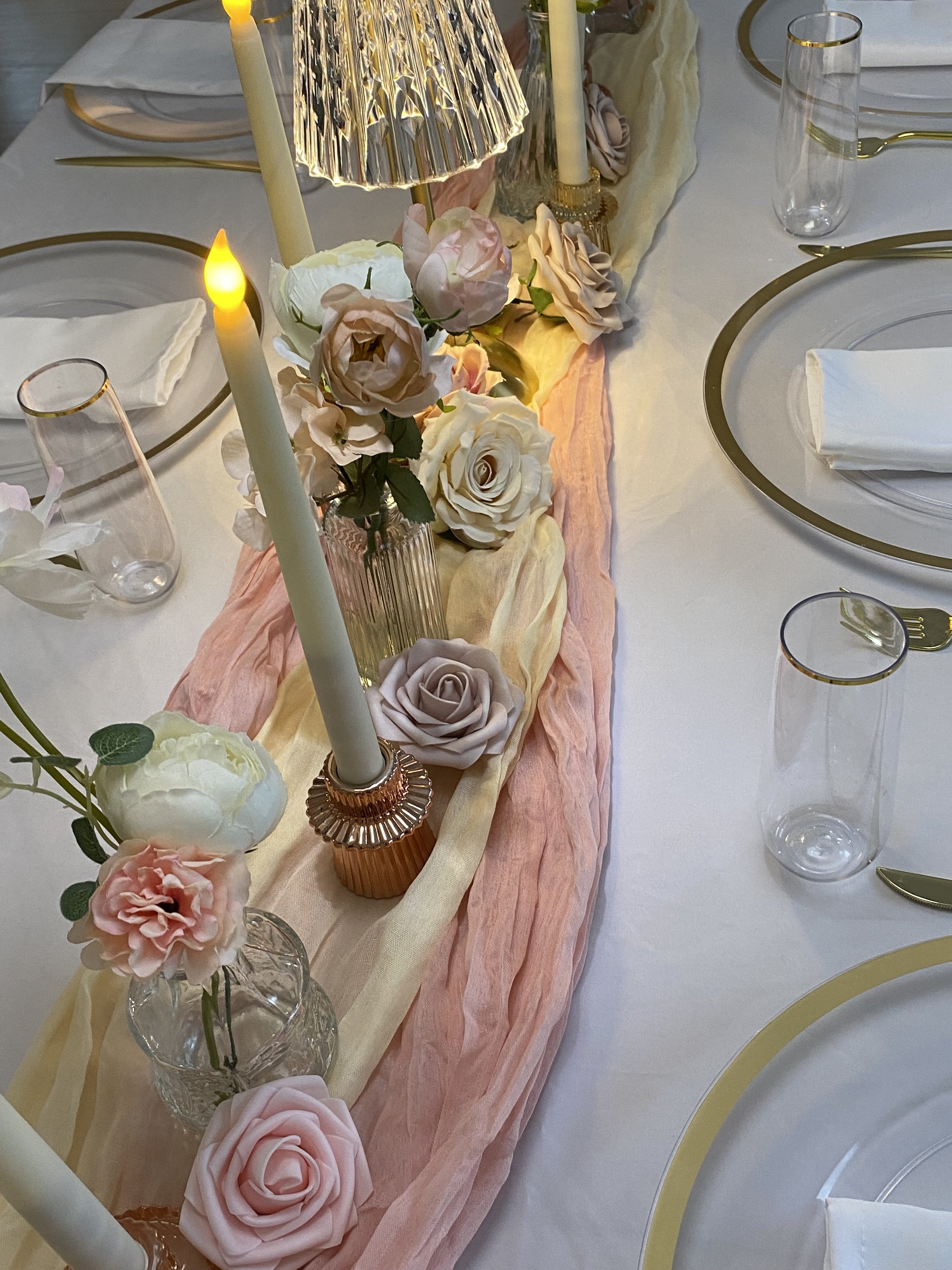 Elegant dining table with floral centerpiece, tall candles, gold-rimmed glassware, and gold-accented tableware, decorated with pastel-colored fabric runner.