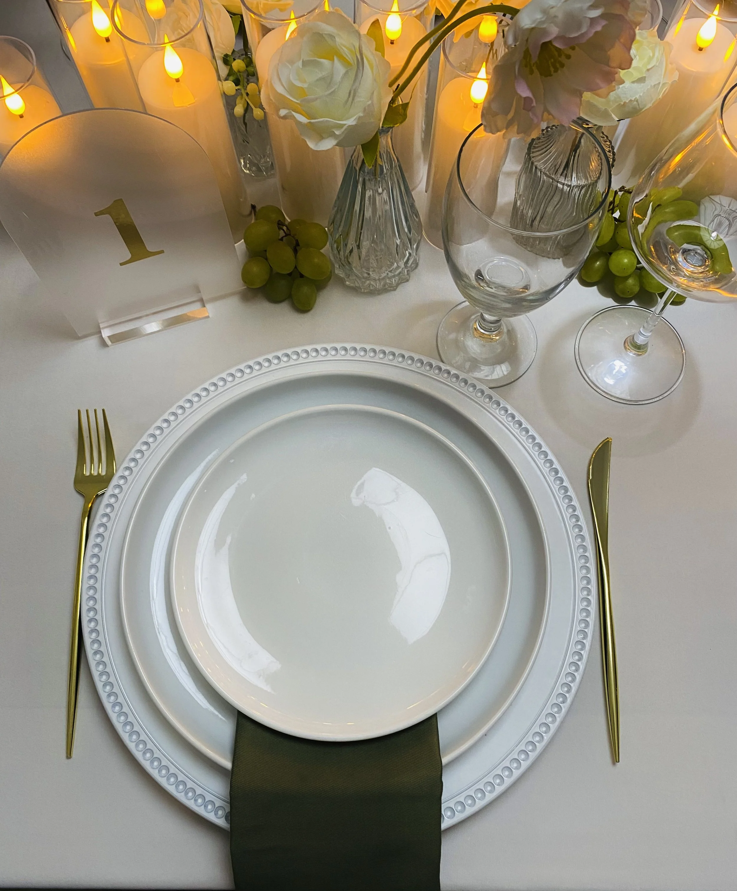 Elegant table setting with white plates, gold utensils, a green napkin, decorative candles with yellow flames, clear glass vases with white and pink flowers, and small bunches of green grapes, all arranged for a formal event.