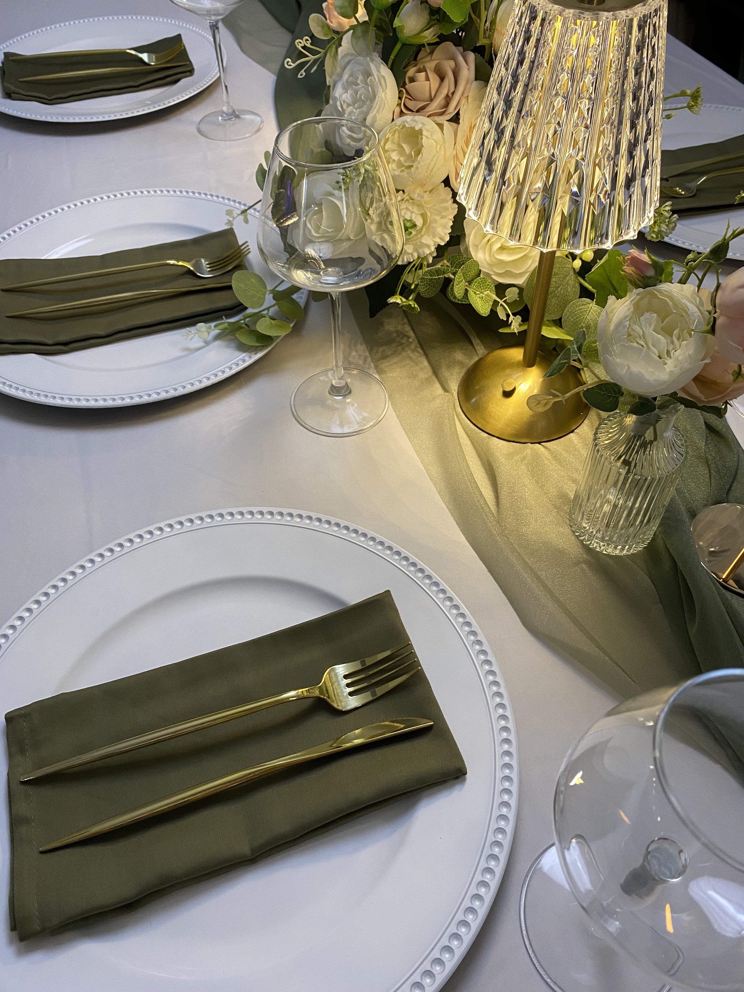 A round white table set for a formal occasion with white plates that have a beaded border, green cloth napkins, gold forks, and empty wine glasses. The center decoration includes a floral arrangement with white and blush roses, white hydrangeas, and greenery. There is a gold and crystal table lamp with a textured shade illuminating the arrangement. An additional clear glass vase with flowers is also visible.