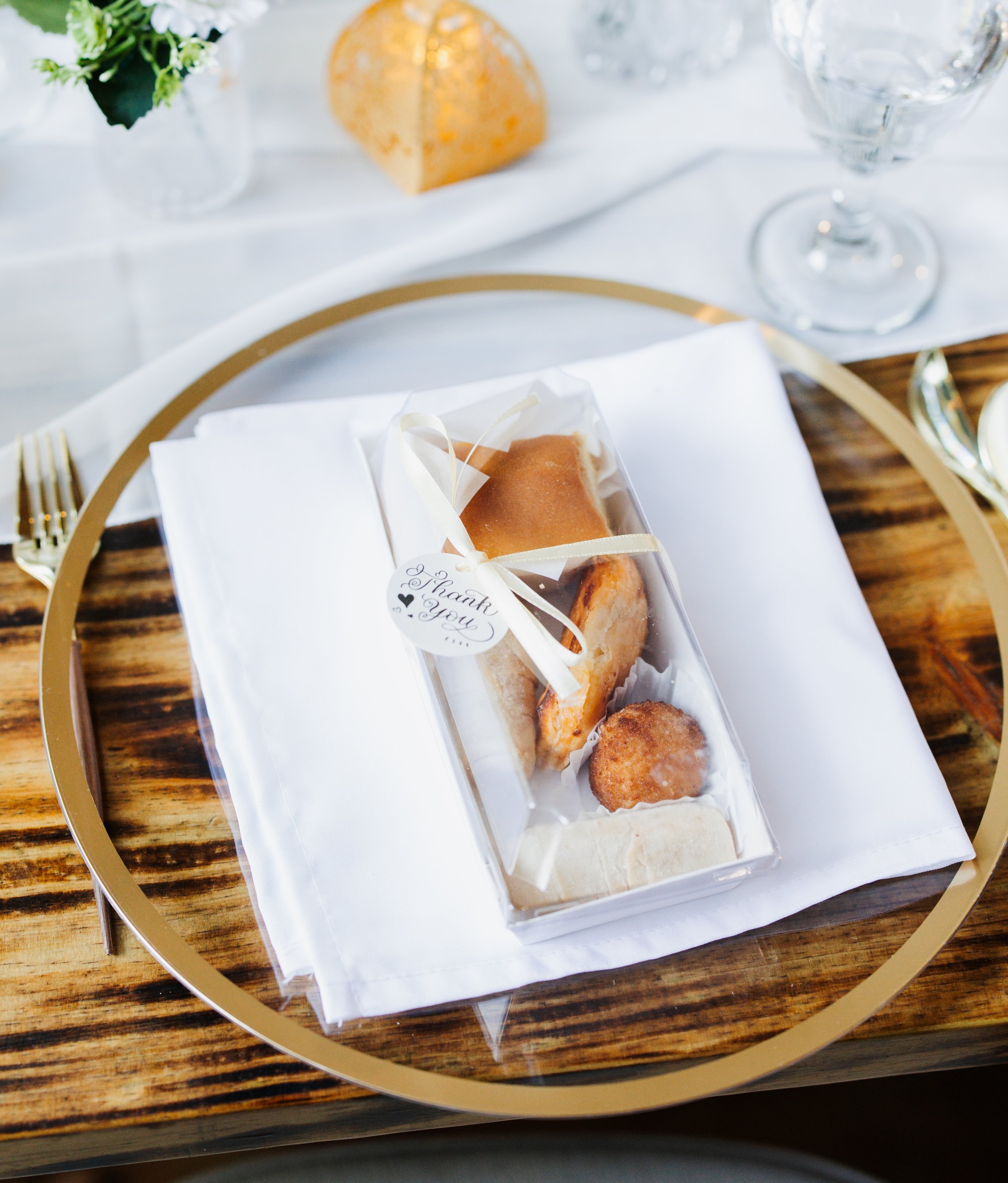 A clear plastic box with assorted pastries, including a glazed pastry, a round pastry with filling, and a crusty breadstick, all on a white cloth napkin on a gold-trimmed plate, with a