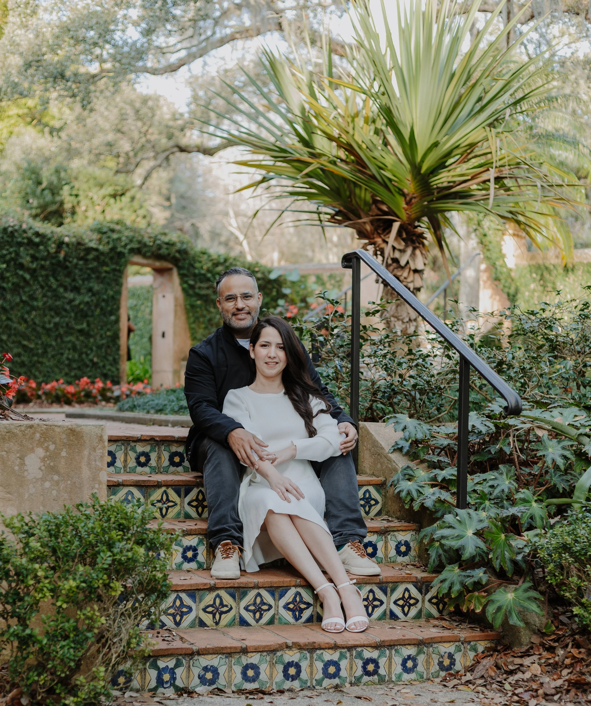 A man and a woman sitting on decorative steps in a garden with lush green plants and trees in the background.