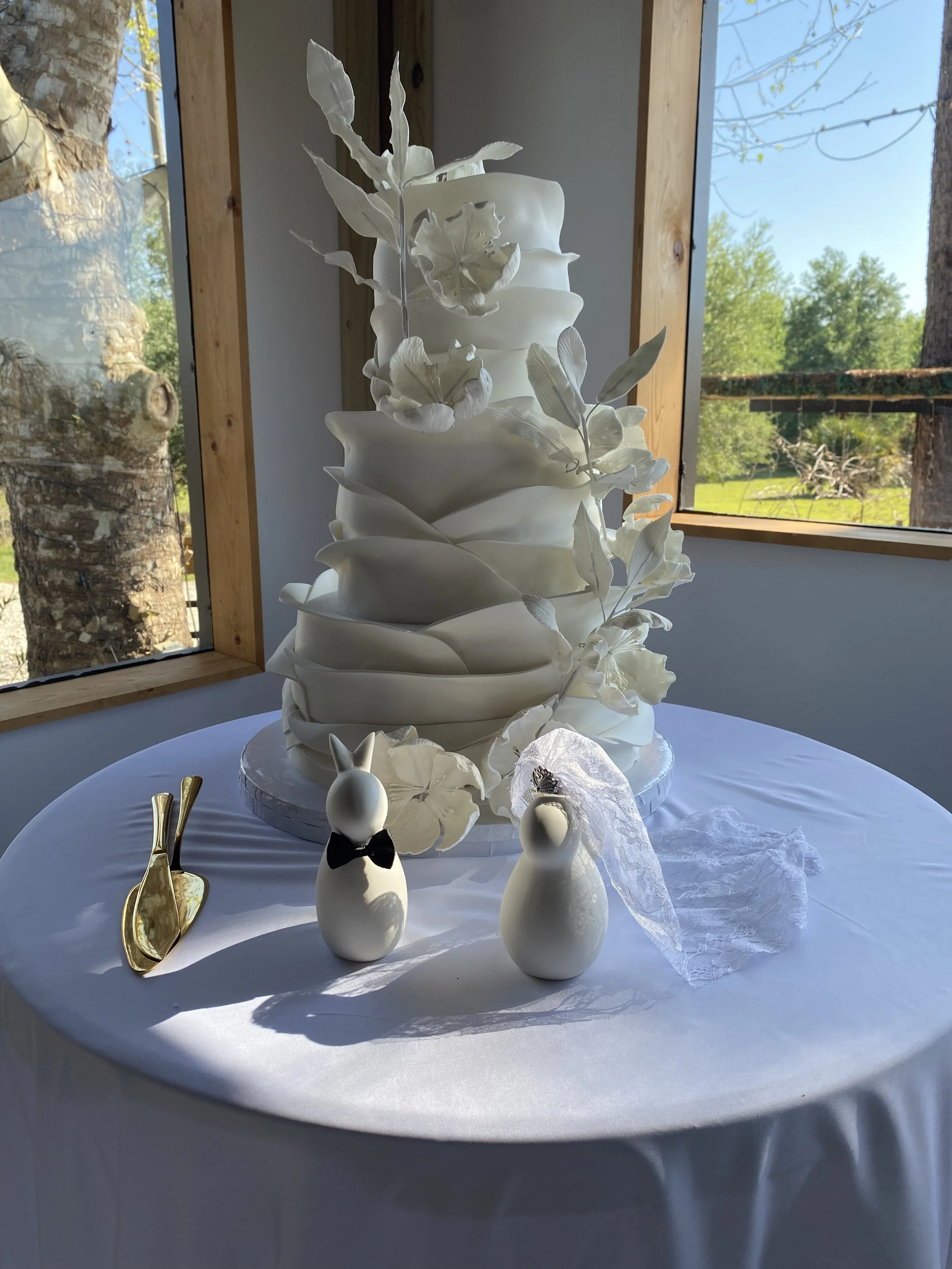 A white tiered wedding cake decorated with white floral and leaf ornaments, placed on a round table with a white tablecloth. Two gold utensils and small decorative white figures are in front of the cake. The background features a window showing green trees and a bright blue sky.