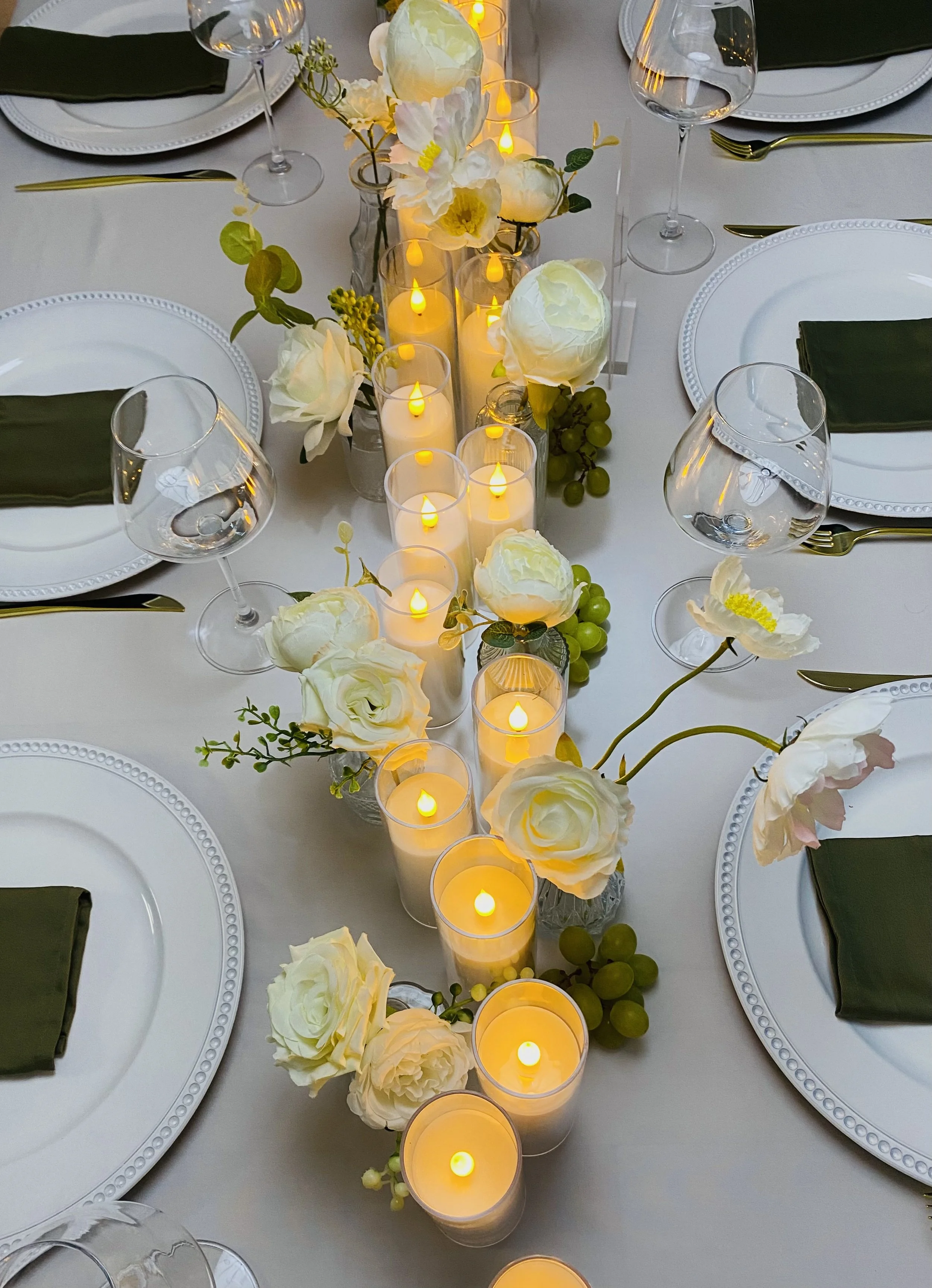 A dining table with white plates, dark green napkins, gold cutlery, wine glasses, and a central floral arrangement with white flowers, green leaves, and small green berries. There are also several candles in glass holders providing soft lighting.