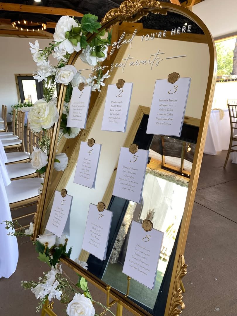 Decorative seating chart with numbered cards and an floral arrangement of white flowers and green leaves. The chart is framed in a gold ornate mirror with the message "We're so glad you're here" at the top.