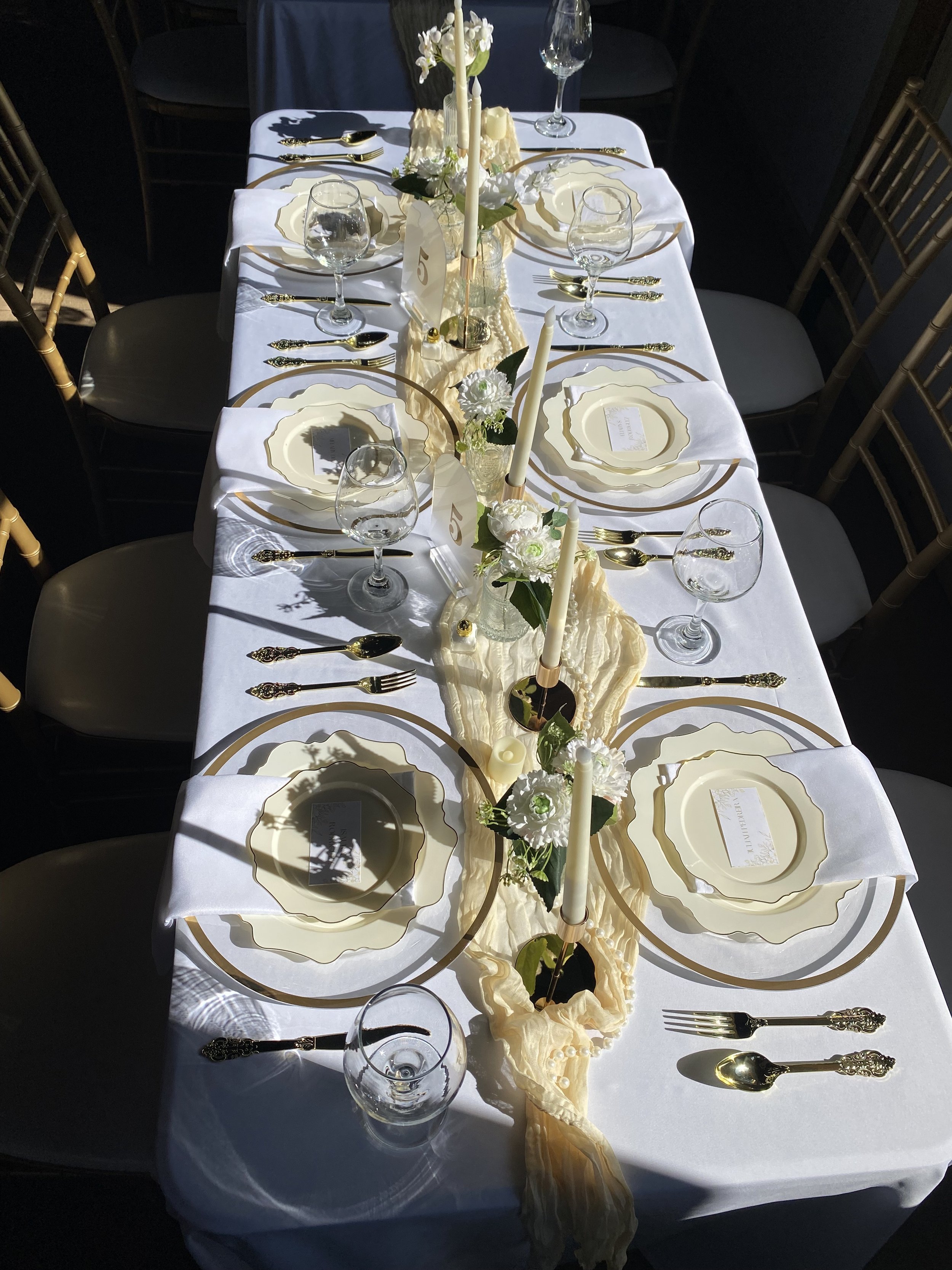 Elegant rectangular dining table set for a formal event with white tablecloth, gold-rimmed plates, gold utensils, wine glasses, floral centerpiece with white flowers and candles, with chairs on both sides.
