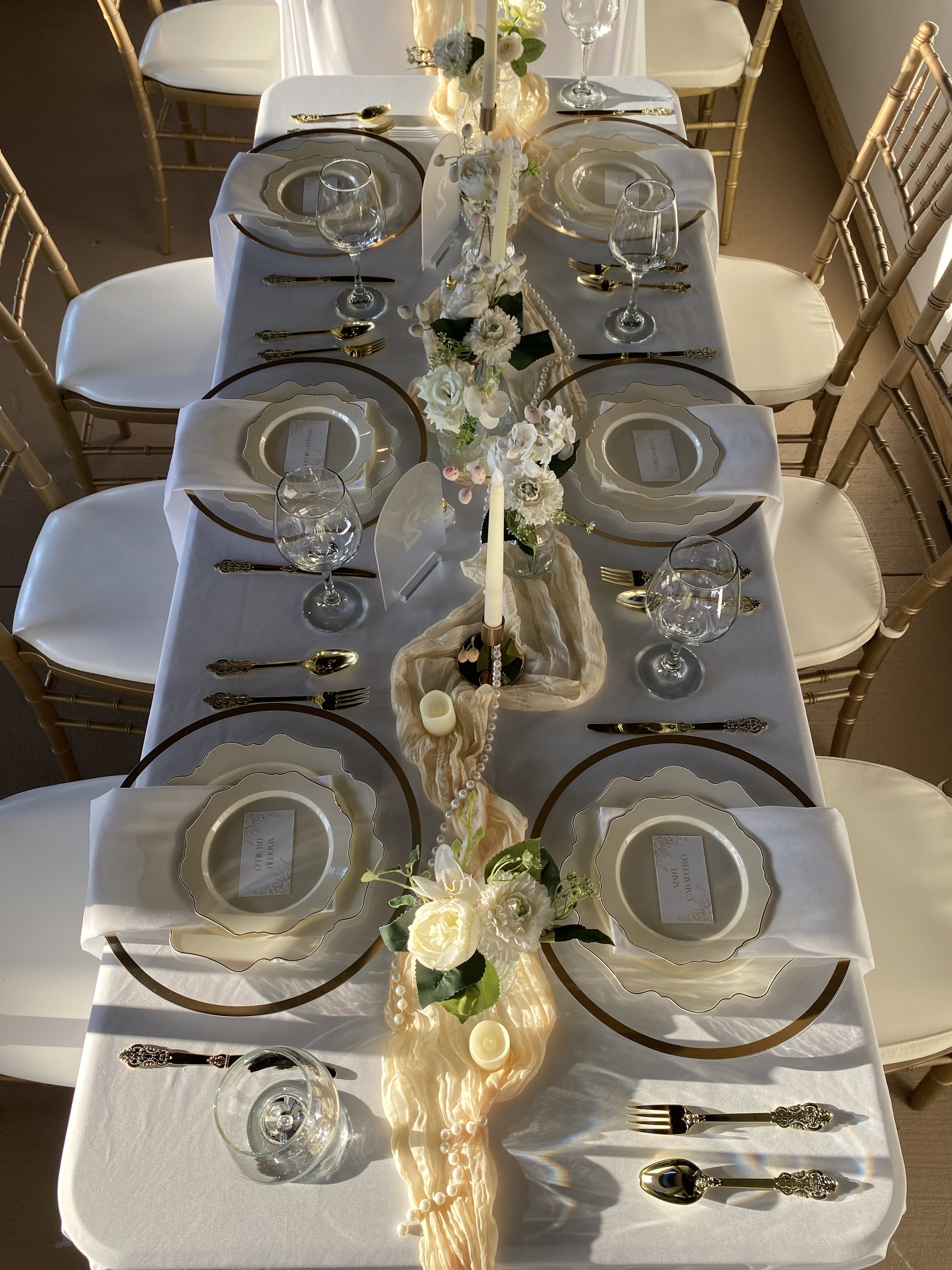 A formal dining table set with white tablecloths, gold-trimmed plates, gold cutlery, wine glasses, and floral centerpiece with white flowers and pearls.