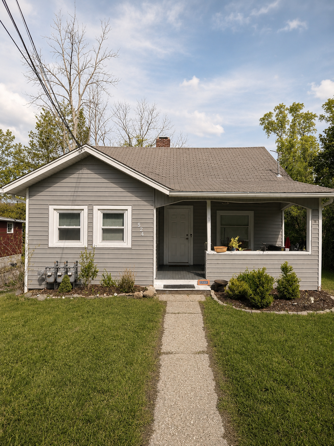 Front view of a gray house with a small front porch, a pathway leading to the front door, green lawn, and some bushes. There are three gas meters on the left side, and leafless trees and green trees in the background.