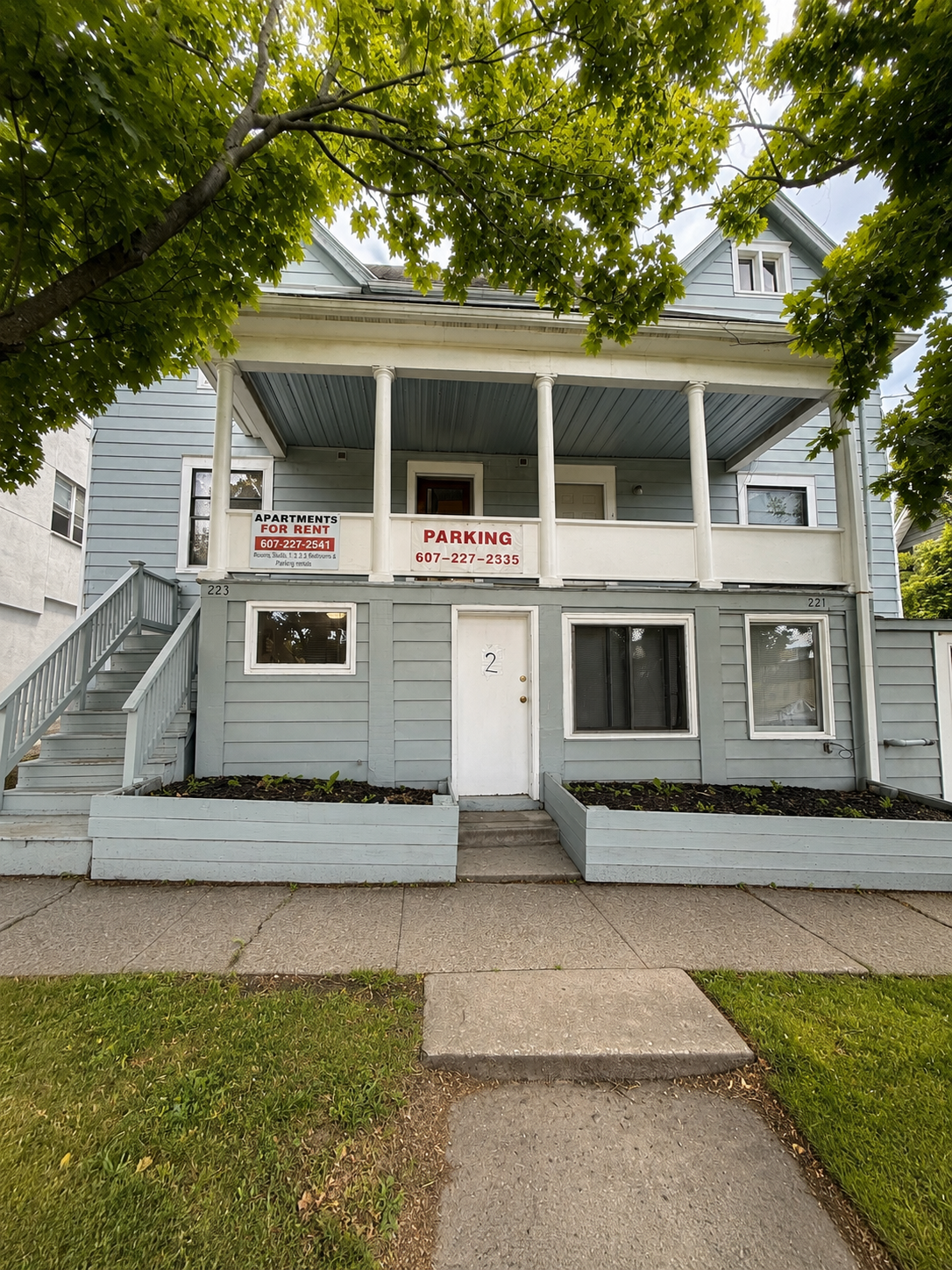 Two-story gray wooden house with a covered front porch, stairs on the left, and garden beds in front. Signs on the porch indicate available apartments for rent and parking contact information. Lush green trees partially frame the house.
