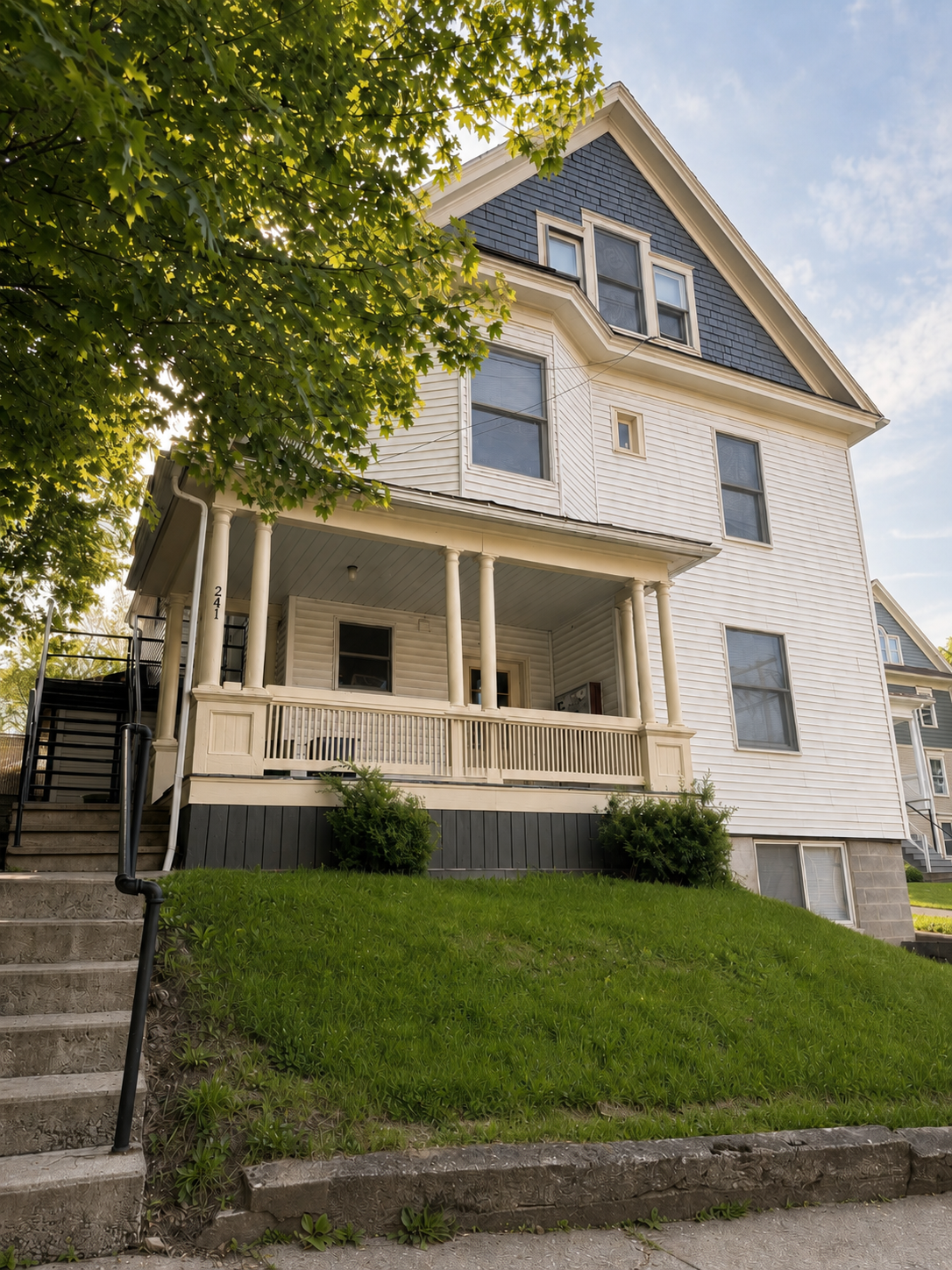 Front view of a three-story white house with a porch and a gable roof, surrounded by a green lawn, with stairs and a tree in the foreground.