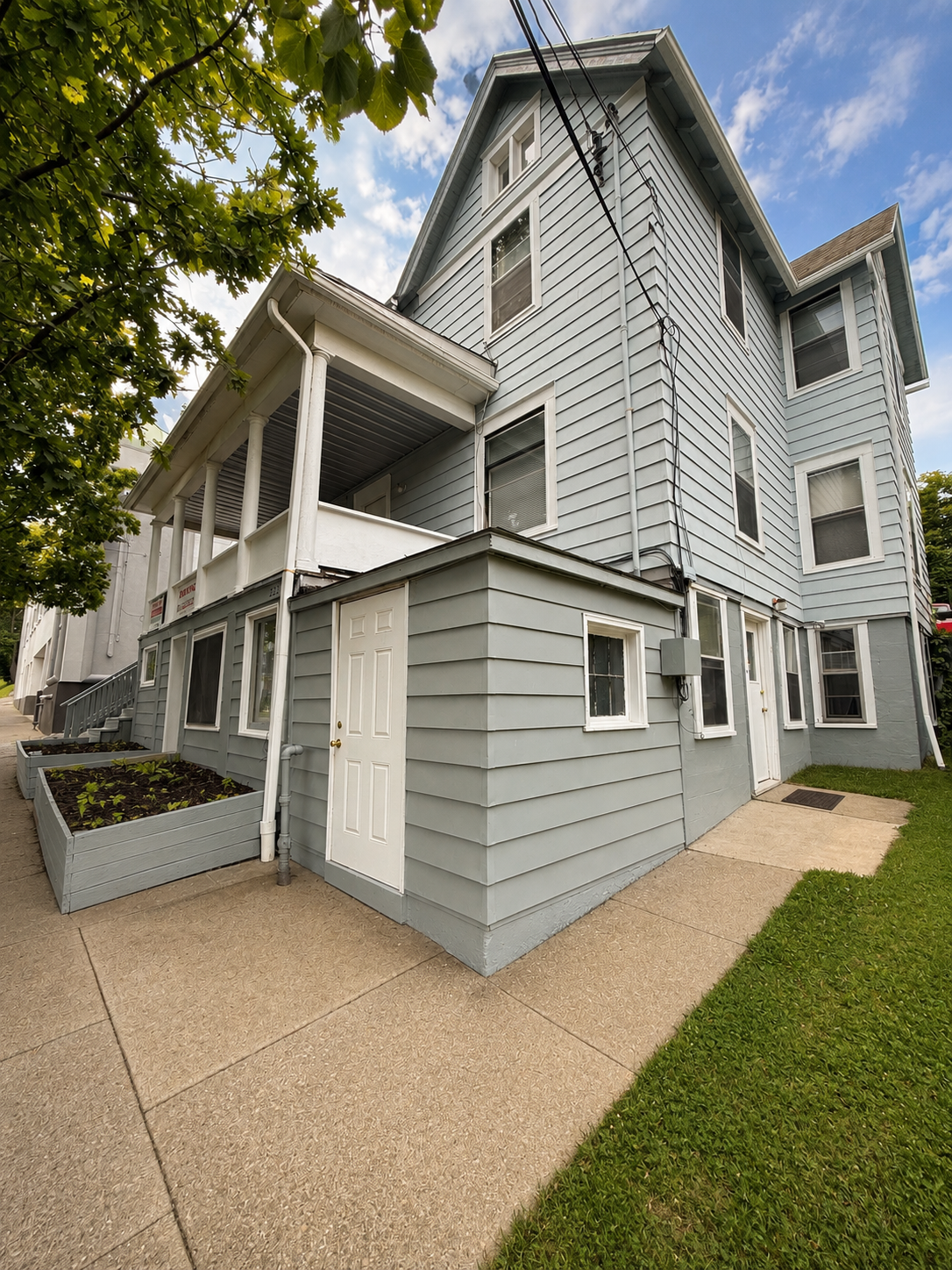 Two-story house painted light blue with multiple windows, a side door, a small porch, and a landscaped garden bed. A sidewalk runs along the house, with green grass and trees nearby, under a partly cloudy sky.