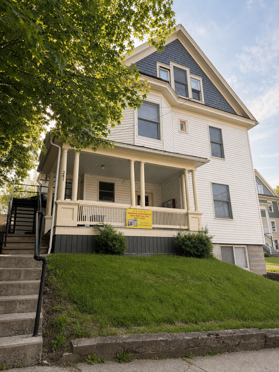 A three-story white house with a dark blue gabled roof, front porch with columns, and stairs leading up to it. There is a yellow sign on the porch advertising apartment rentals.