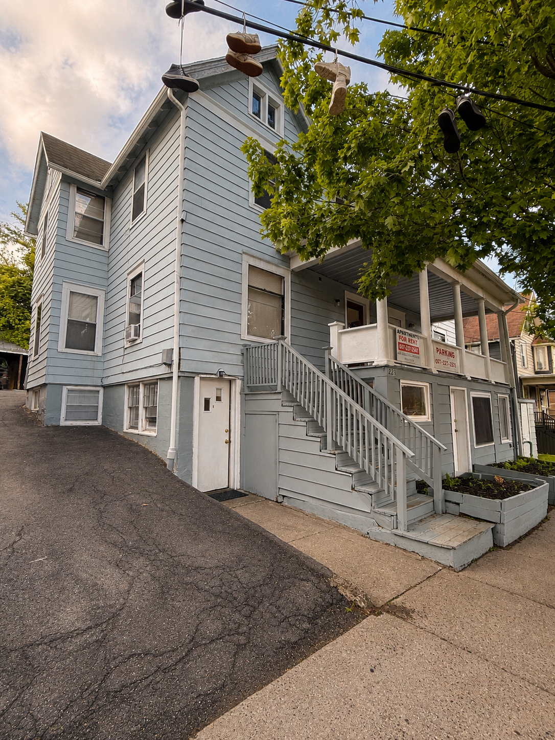 A two-story light blue house with white trim, a staircase leading to the porch, and a 'For Rent' sign on the porch. Shoes hanging on a wire above the sidewalk with greenery and cloudy sky in the background.