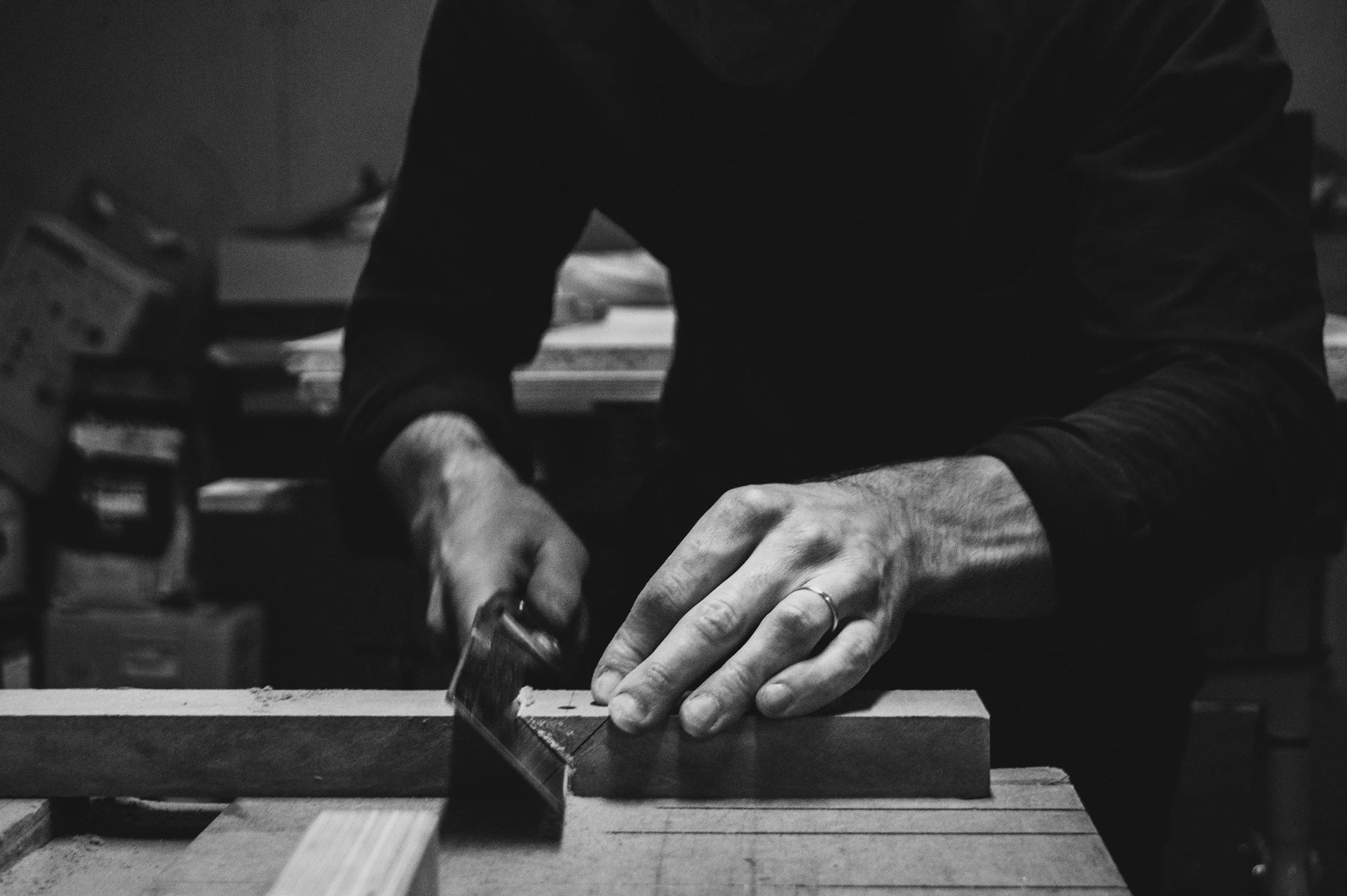 Black and white photo of a person working with a handsaw on a piece of wood, wearing dark clothing, in a workshop or woodworking space.