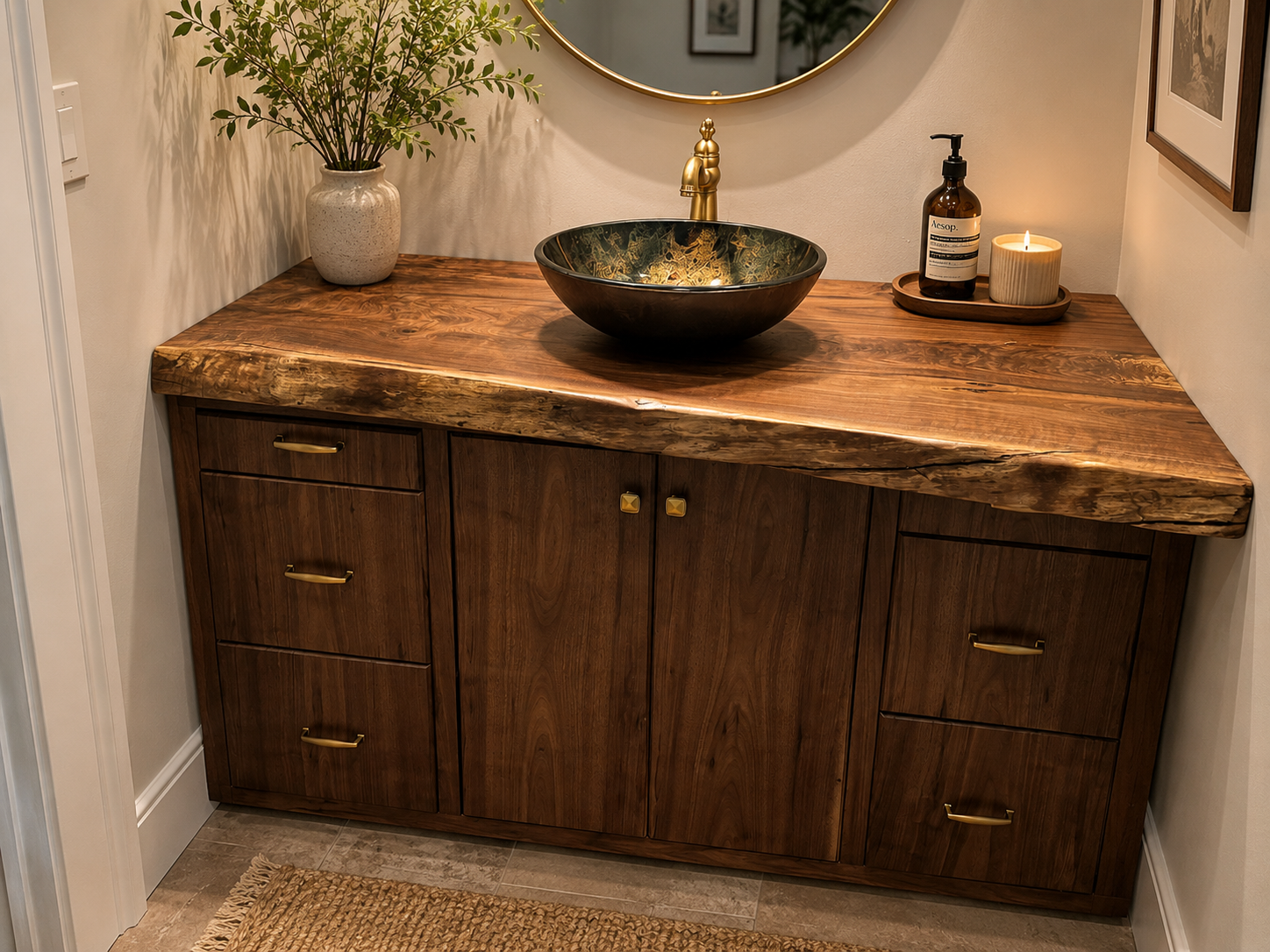 Wooden bathroom vanity with a live edge countertop, a black and gold vessel sink, and a gold faucet. Decor includes a potted plant, a candle, and a soap dispenser, with framed art on the walls.