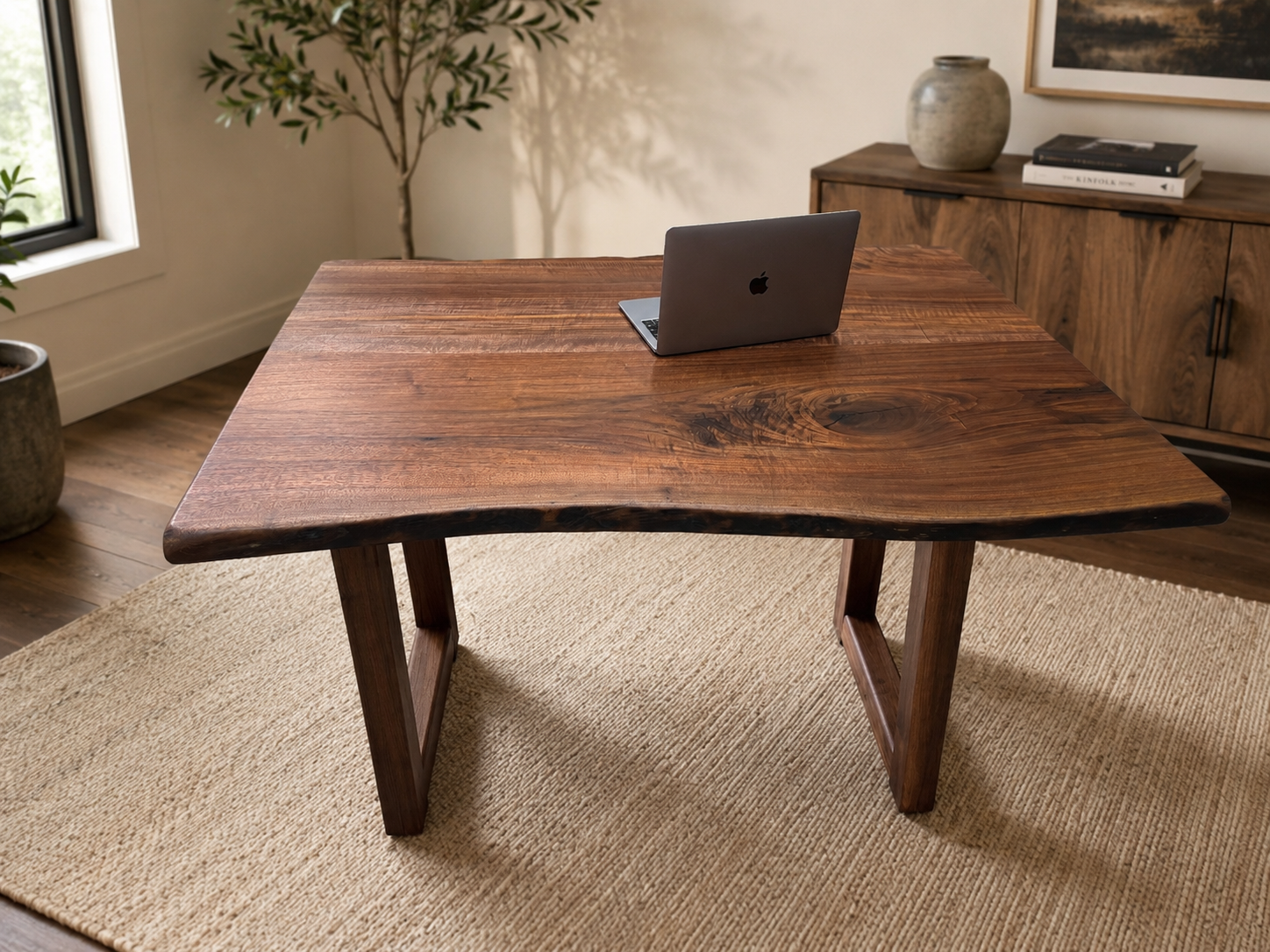 A wooden table with a MacBook laptop on it in a room with a beige rug, a window, a potted plant, and a wooden sideboard with decorative objects.