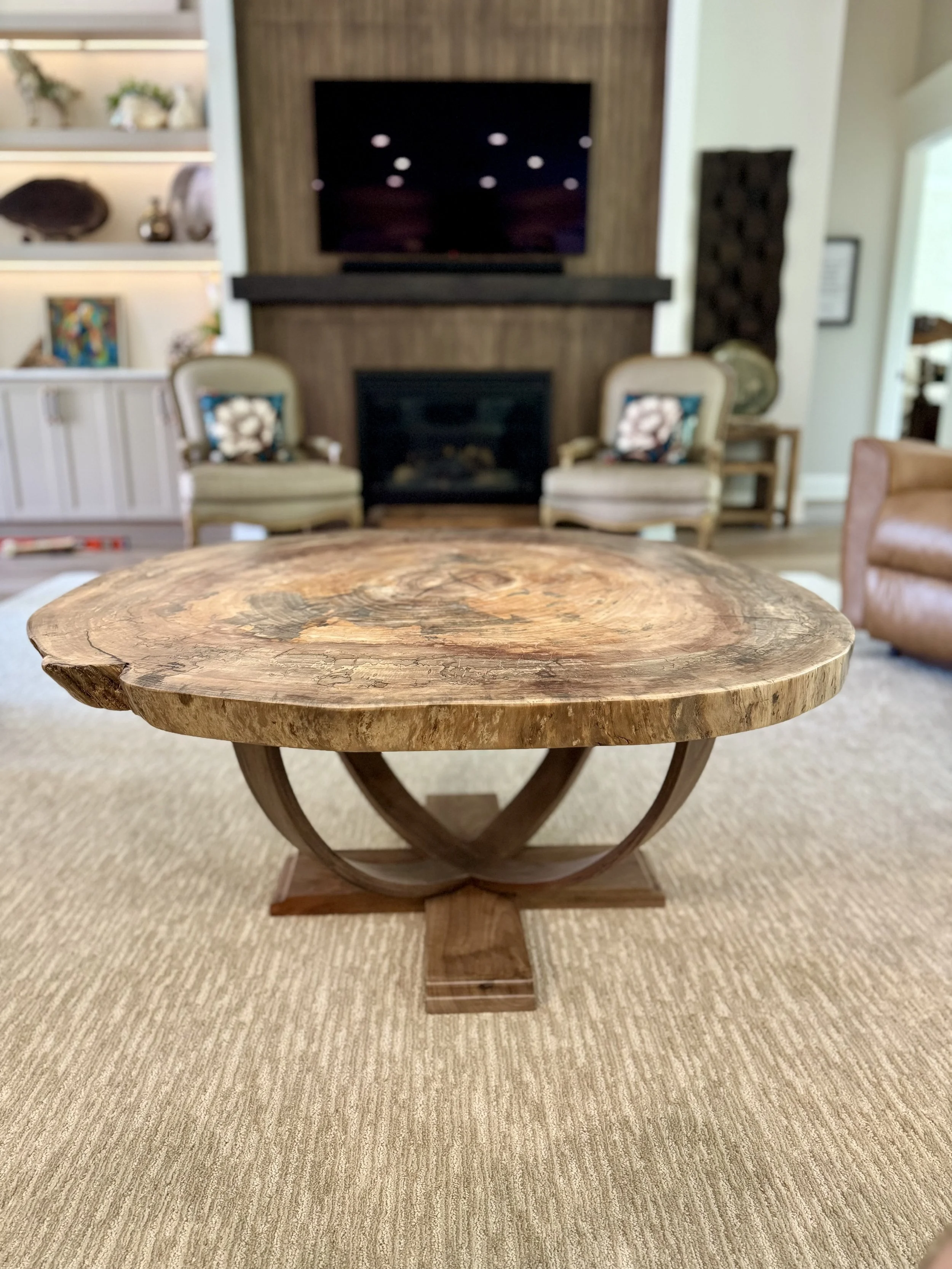 Close-up of a wooden side table on a beige carpet, in a living room with seating and a fireplace in the background.