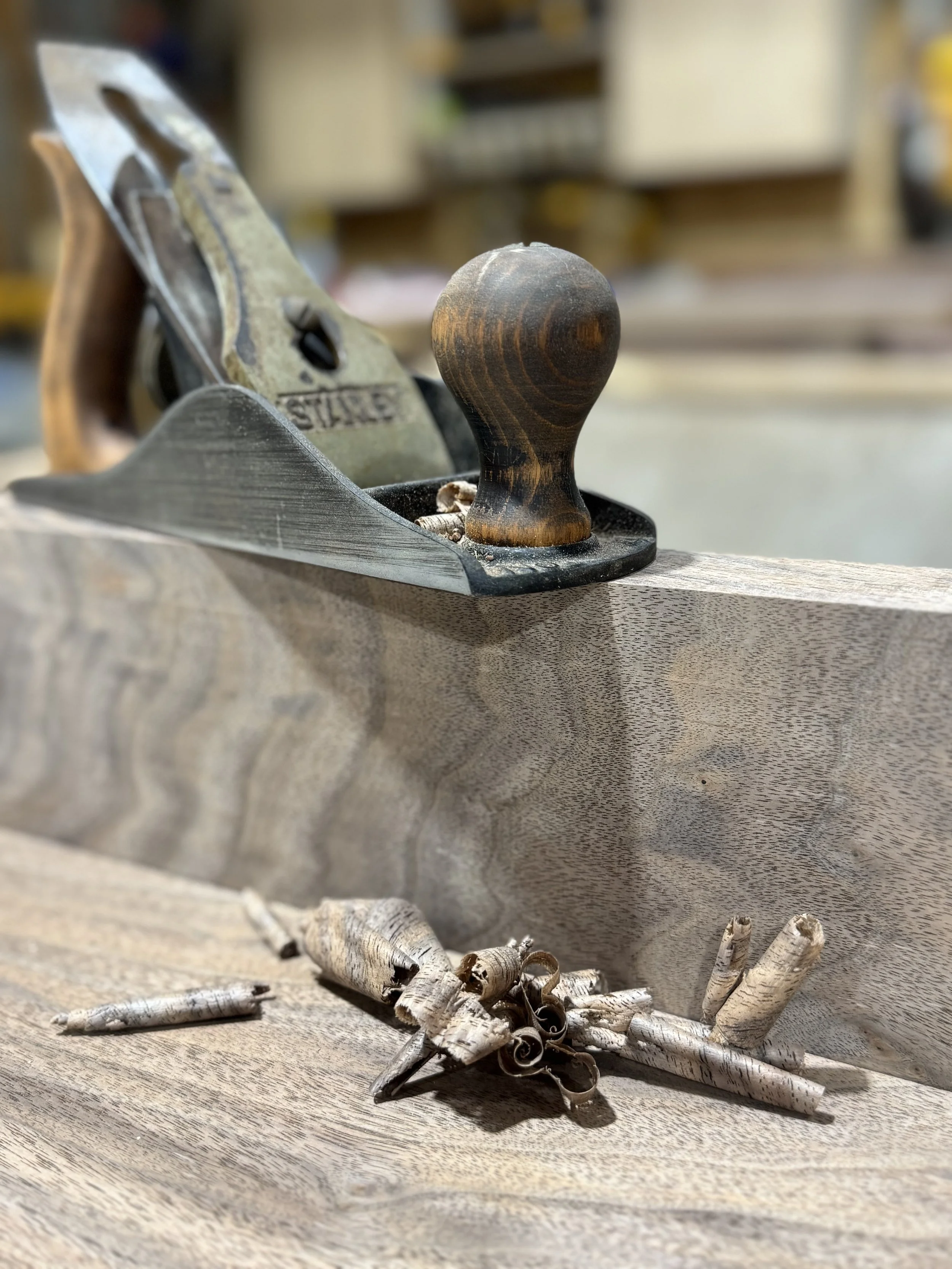 A woodworking hand plane, a wooden-handled tool with a metal body, rests on a wooden work surface beside a bundle of dried birch bark.