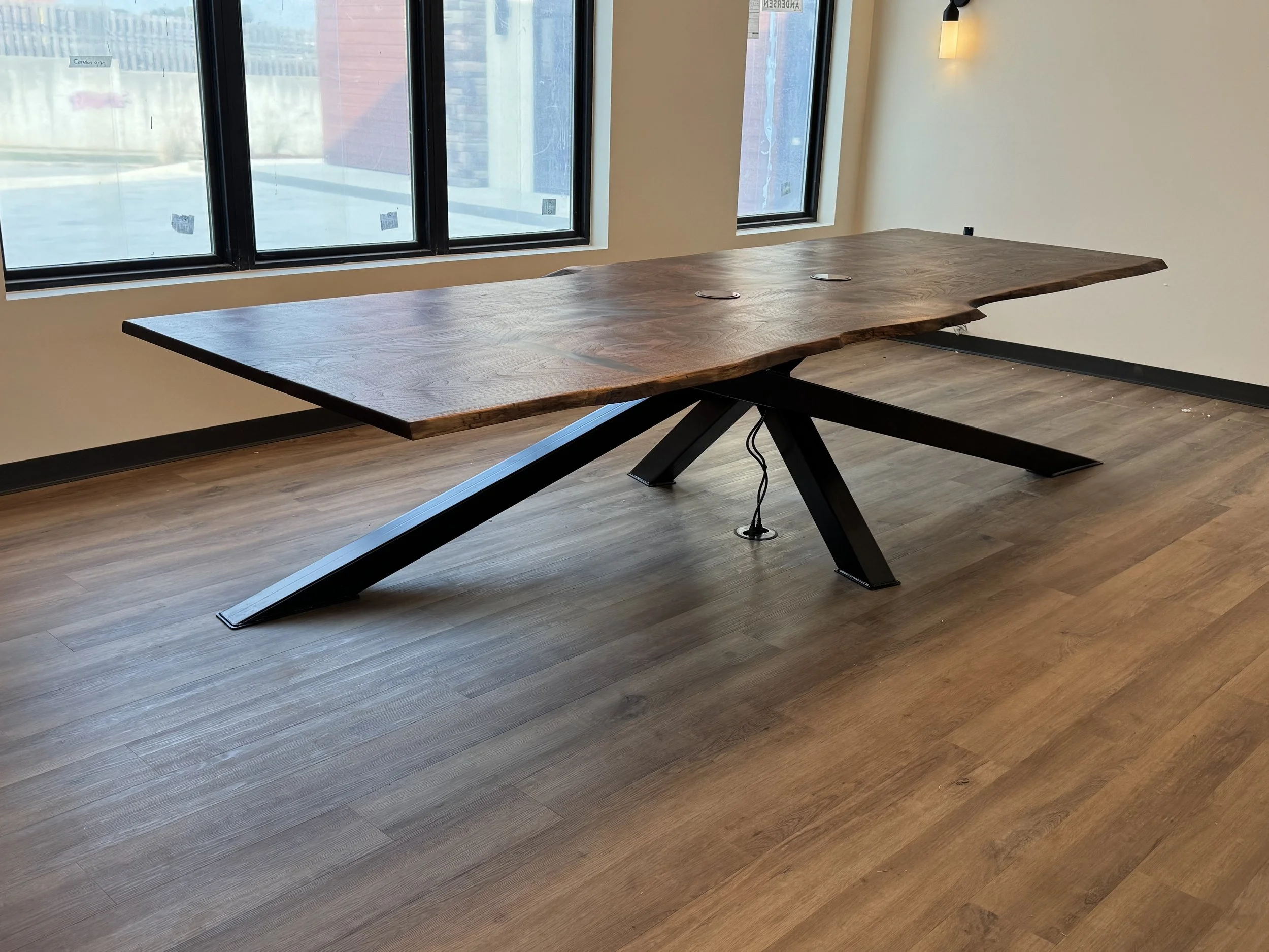 A wooden table with a polished, natural edge, supported by black metal legs, placed next to large windows with a view of building exteriors and a concrete outdoor area, and illuminated by a wall sconce light.