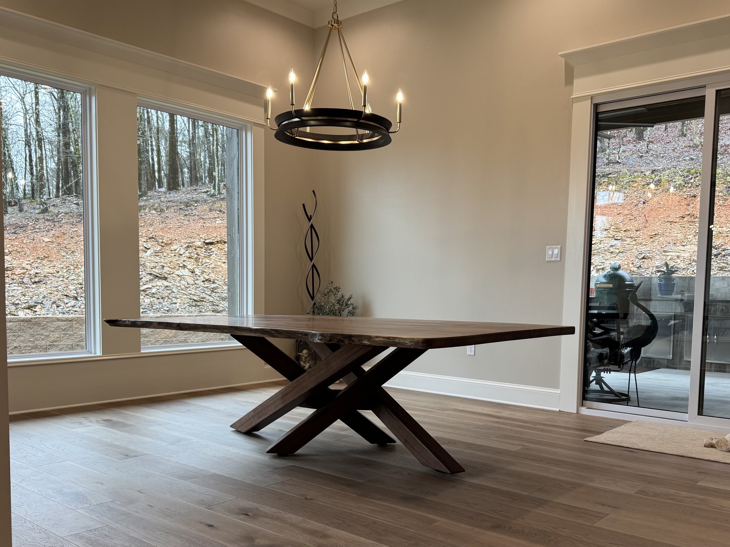 Interior of a room with large windows showing an outdoor wooded landscape, a modern chandelier, a wooden dining table, and a sliding glass door leading to an outdoor patio.