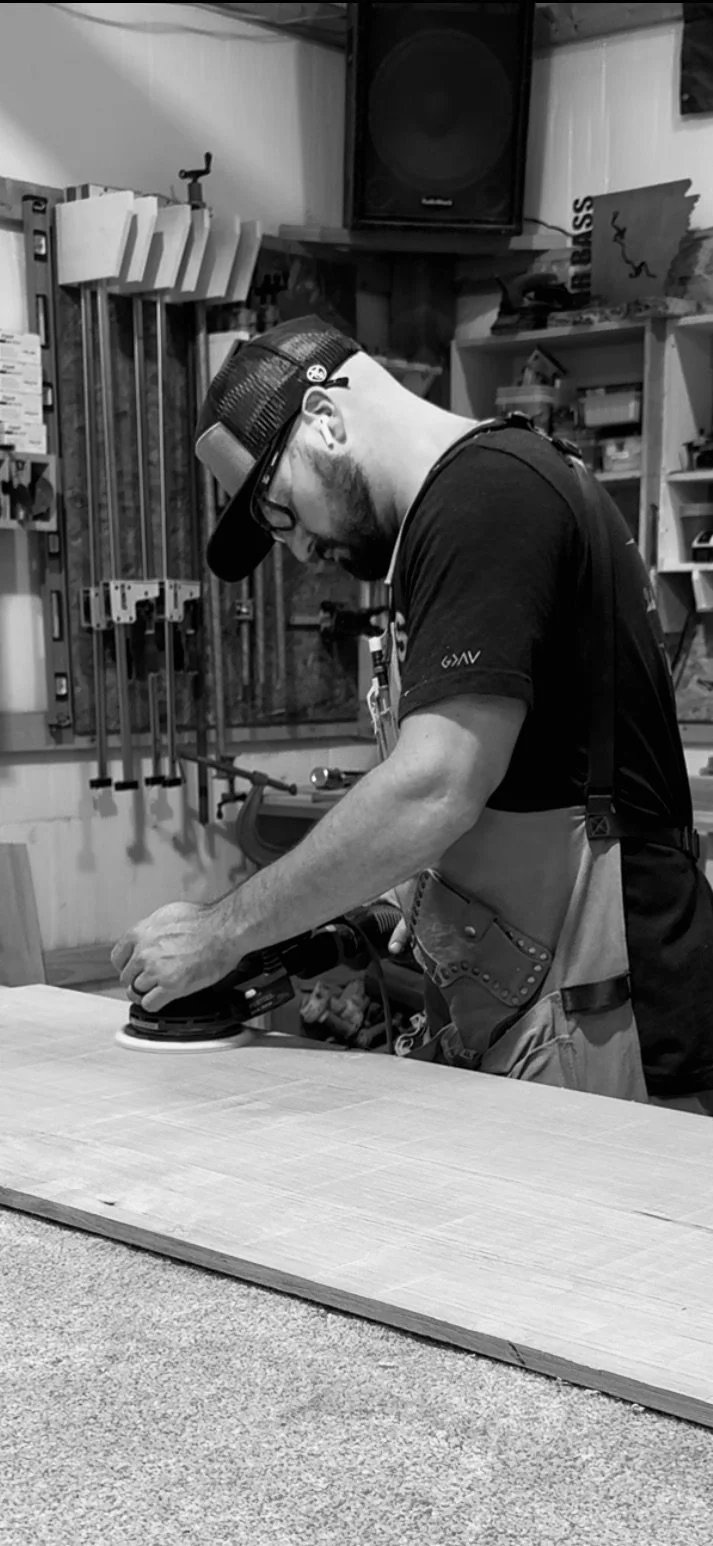 A man sanding a piece of wood with a power sander in a workshop.