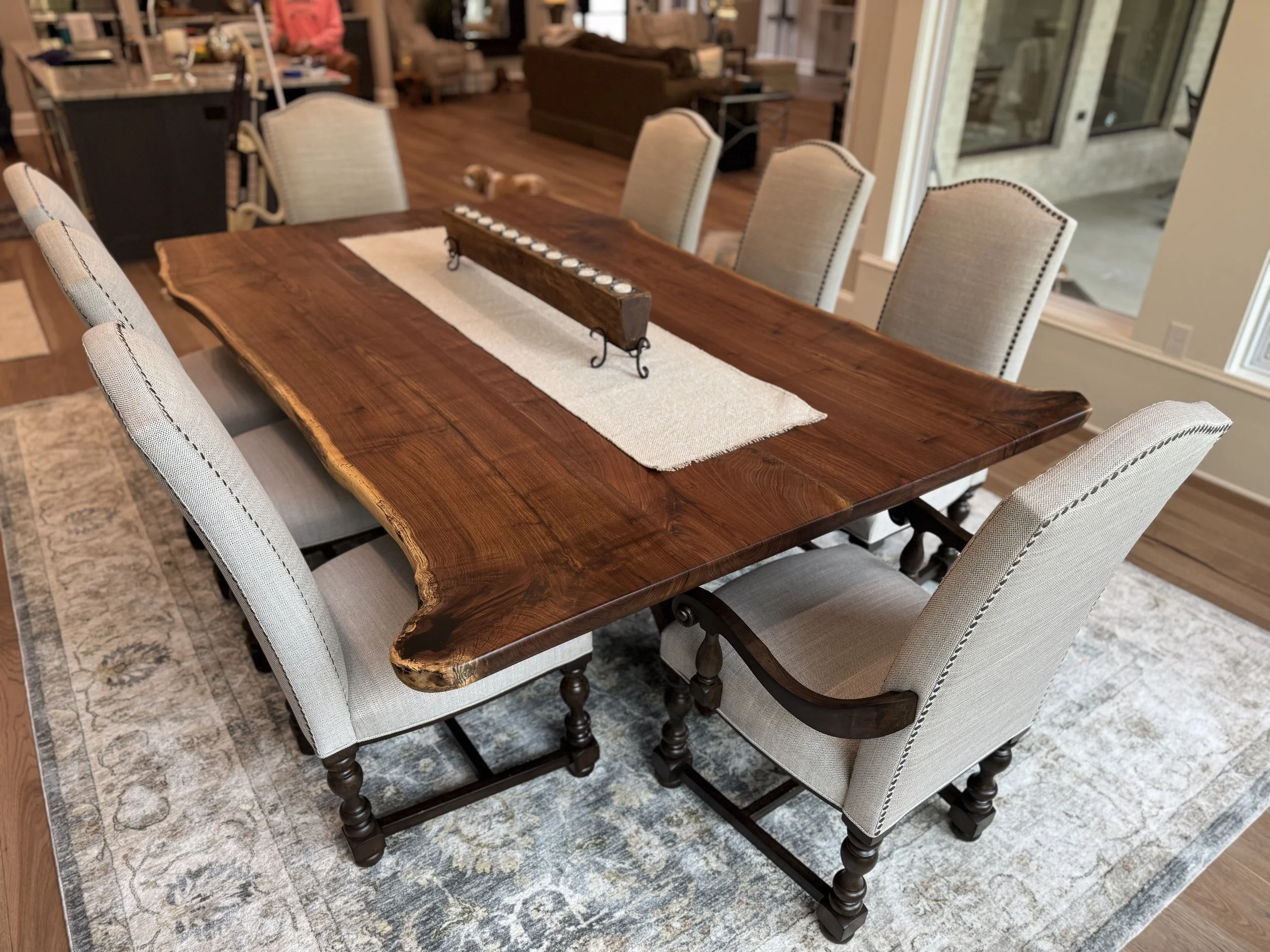 A large wooden dining table with a natural edge and a beige table runner, surrounded by eight upholstered dining chairs with light fabric and dark nailhead trim, in a spacious open-concept living and dining area.