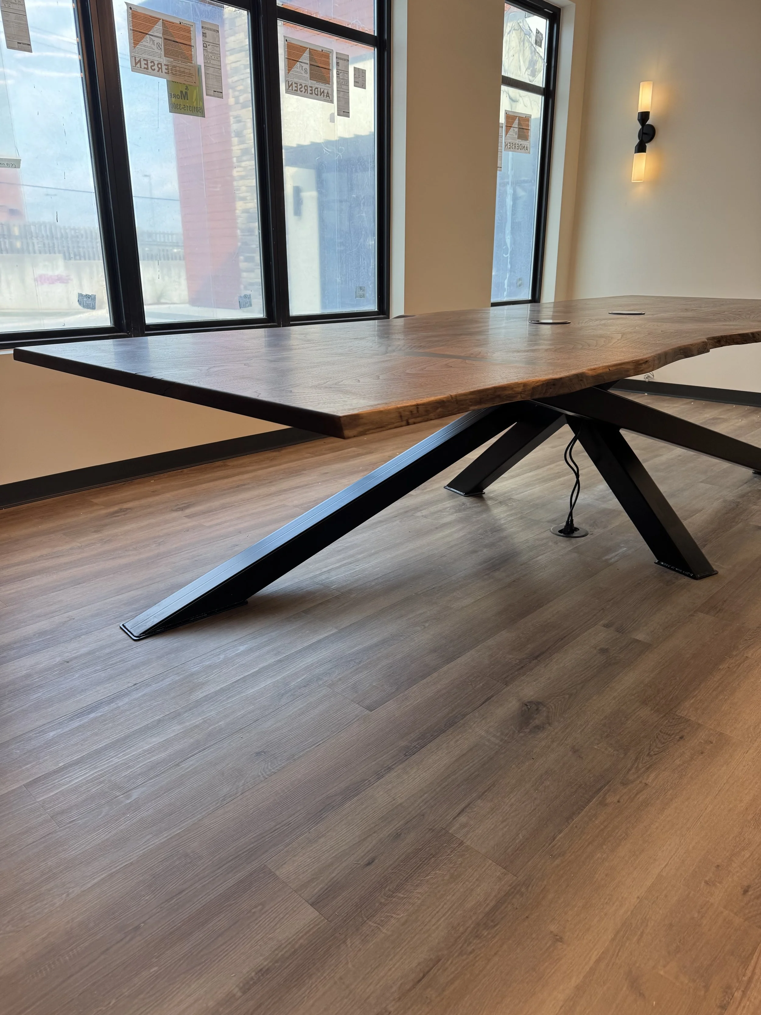 A large, rustic wooden table with black metal legs in a room with wood flooring and large windows, with printed notices taped to the windows and the wall light turned on.