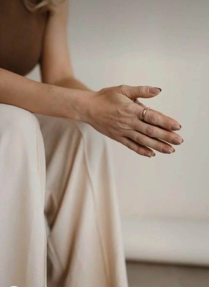 A close-up of a woman's hands resting on her lap. She is wearing a ring on her finger and a beige or light-colored dress.