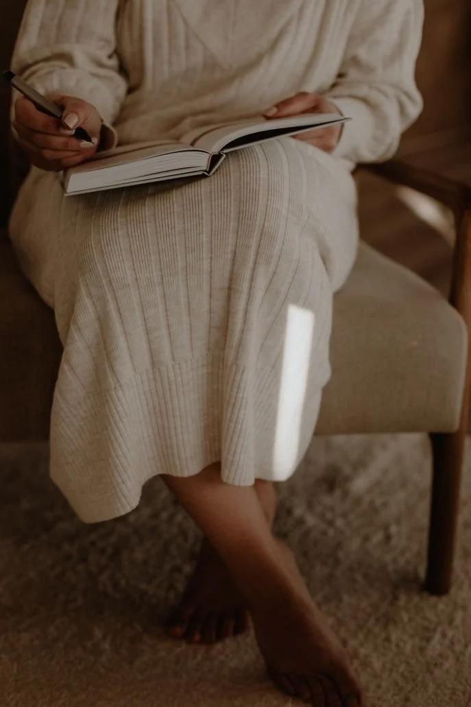 Person sitting on a beige chair, wearing a cream-colored sweater and skirt, holding an open notebook and a pen, with crossed legs and barefoot on a carpeted floor.