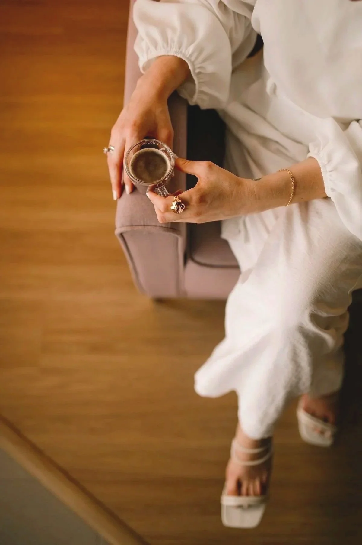 A woman wearing off-white clothing holding a cup of coffee while sitting on a beige sofa in a room with wooden flooring.